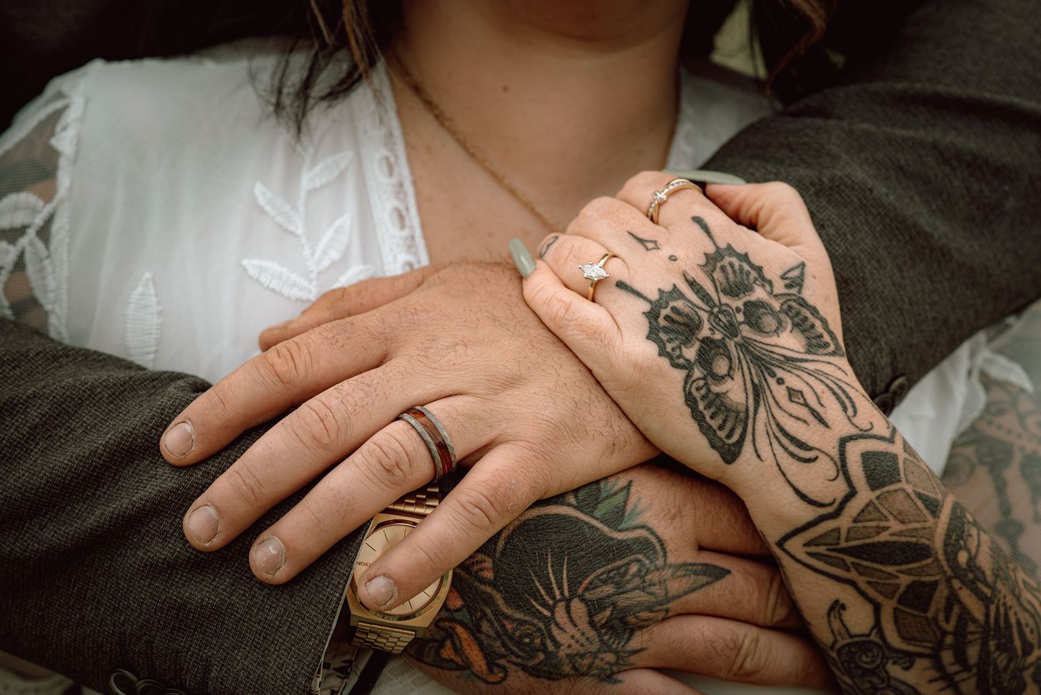 Close-up of couple's hands showing wedding rings, with bride's tattooed arms wrapped around groom's suit jacket.