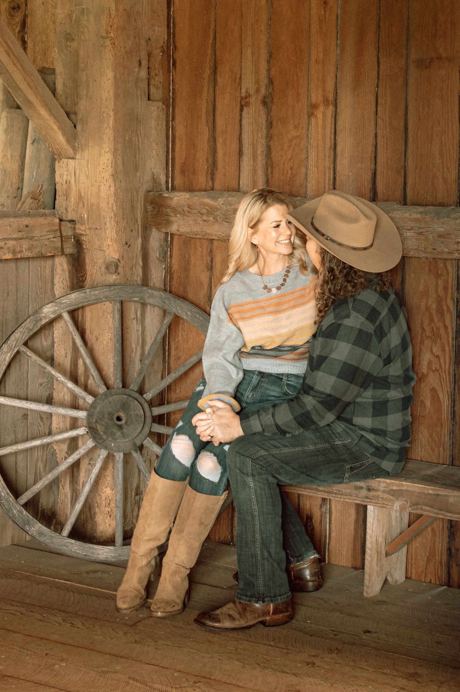 Couple in Western attire sitting on a bench in a rustic barn, with a wooden wagon wheel backdrop.