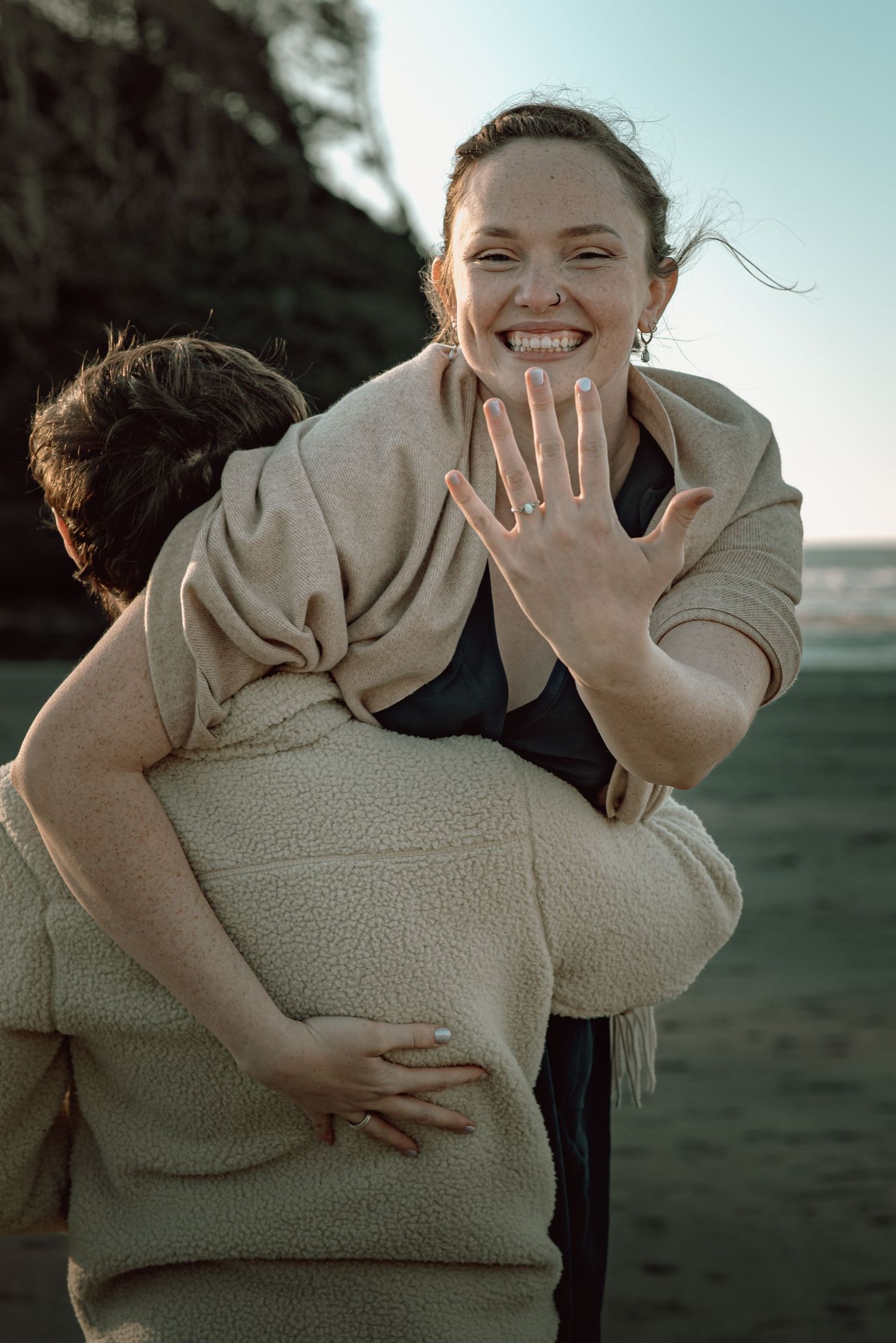 Excited newly engaged woman showing ring while hugging partner on beach.