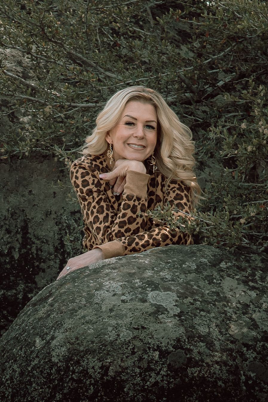 Smiling blonde woman in leopard print dress poses on a mossy rock surrounded by greenery.