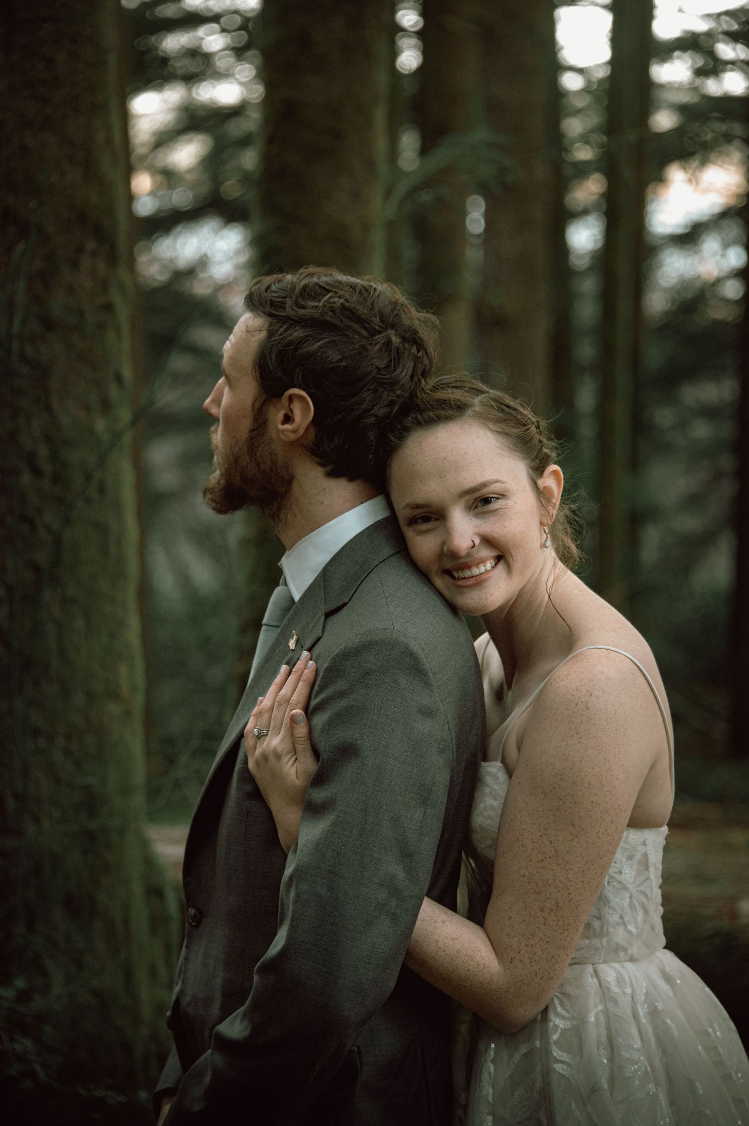 Bride in lace gown leaning against grooms back smiling in tall evergreen forest setting.