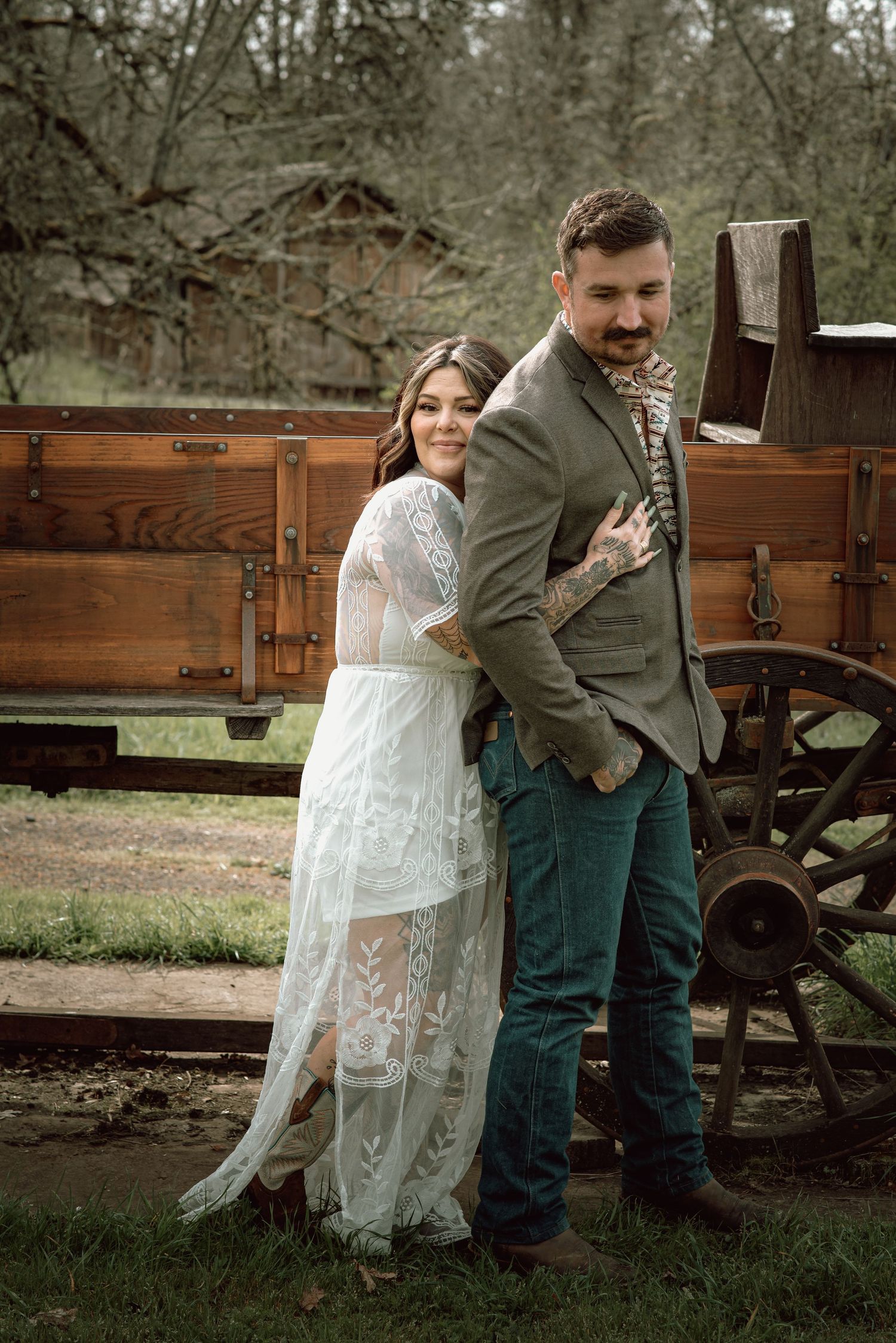 Bride in sheer lace gown leans against groom in olive suit near vintage wooden wagon in outdoor setting.