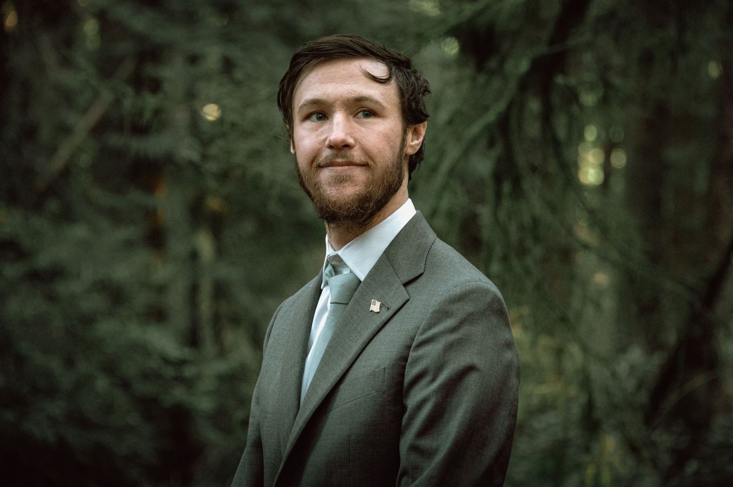 Bearded groom in gray suit with light blue tie standing in a green forest for a wedding portrait.