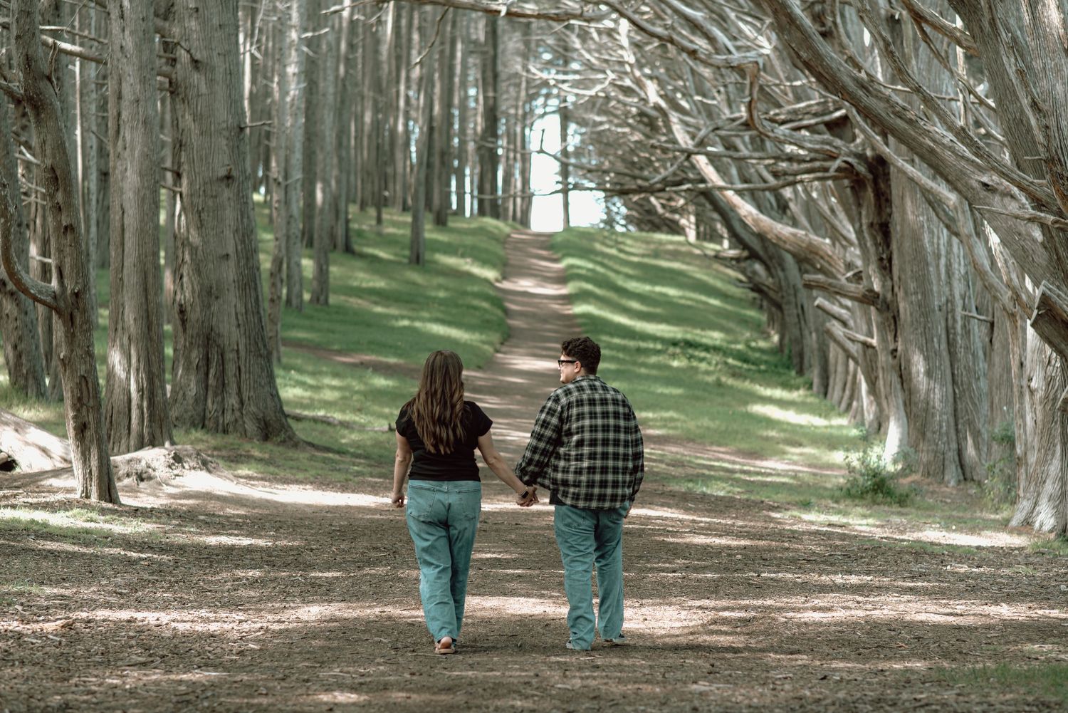 Couple walking hand in hand down a peaceful tree-lined path surrounded by tall trees and green grass.