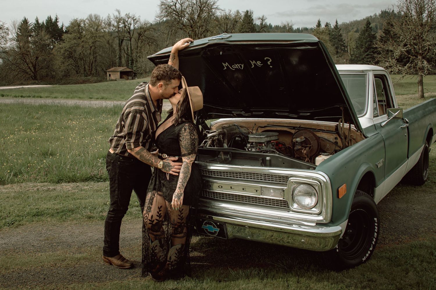 Couple sharing a kiss beside an open hood of a vintage teal truck with Marry Me written inside.