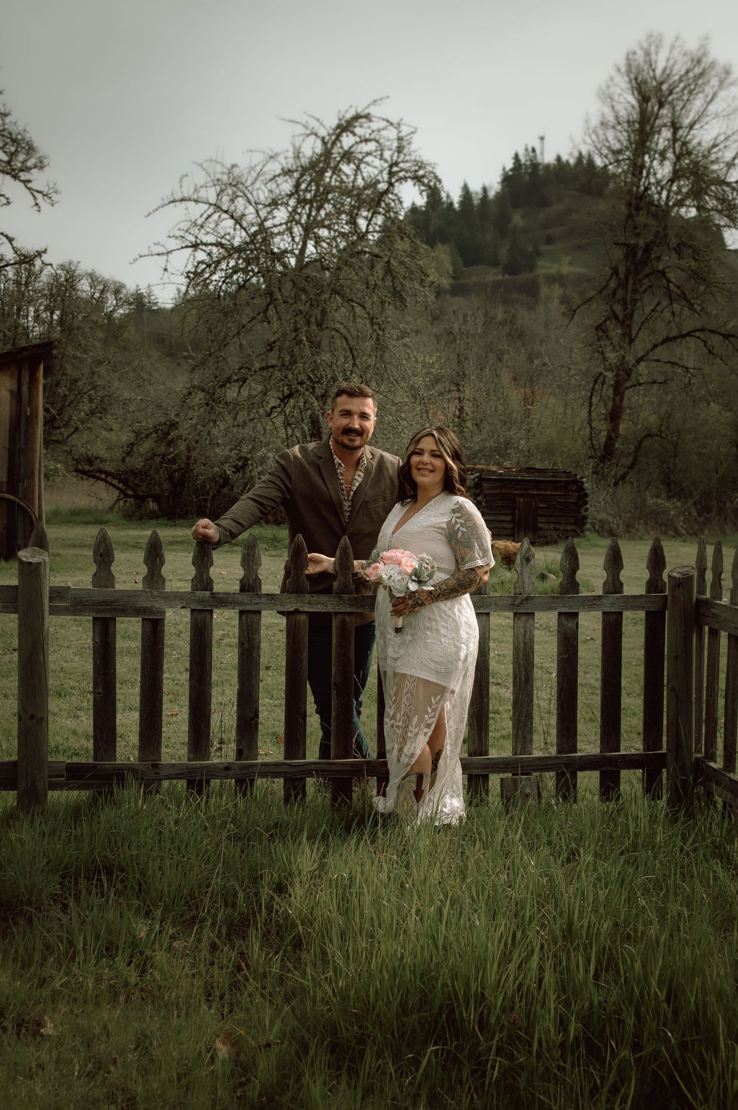 Smiling bride and groom posing by a weathered wooden fence holding a pink bouquet.