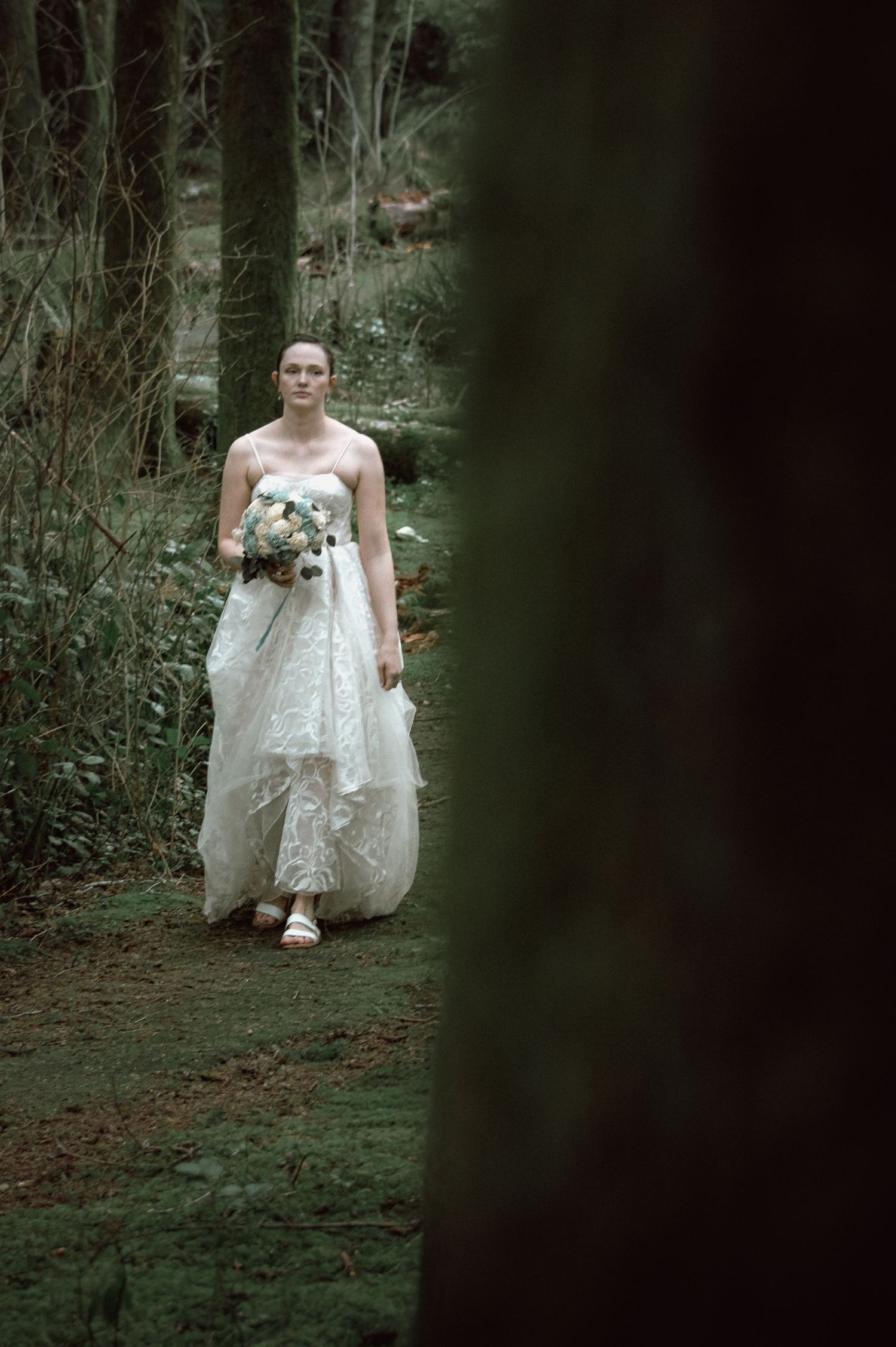 Bride in flowing white lace gown walking through a forest, carrying a bouquet, framed by dark tree trunks.