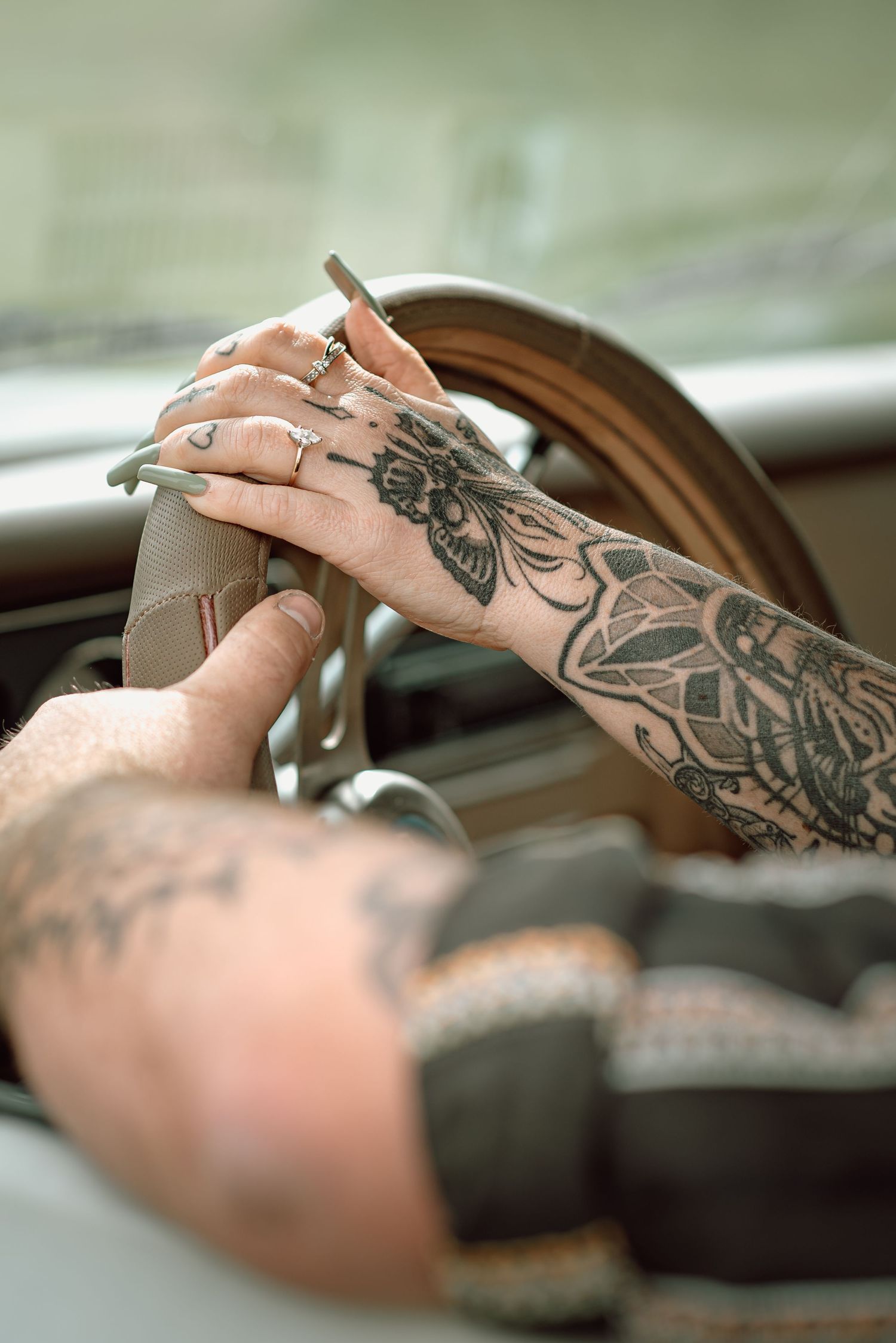 Close-up of a couple holding hands over a vintage steering wheel, both with intricate tattoos and rings.