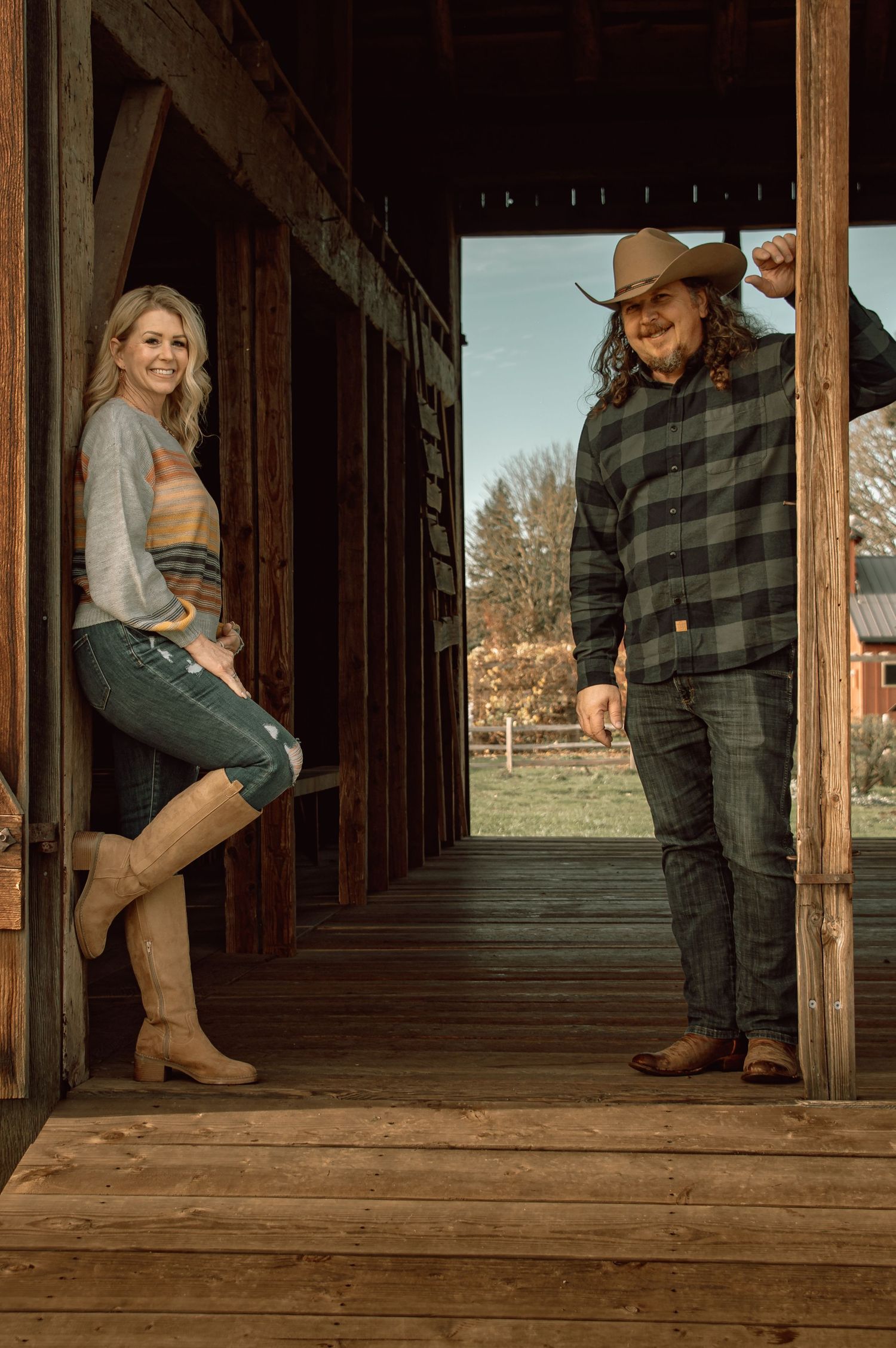 Country couple posing in a rustic barn doorway, woman in boots and man in cowboy hat and plaid shirt.