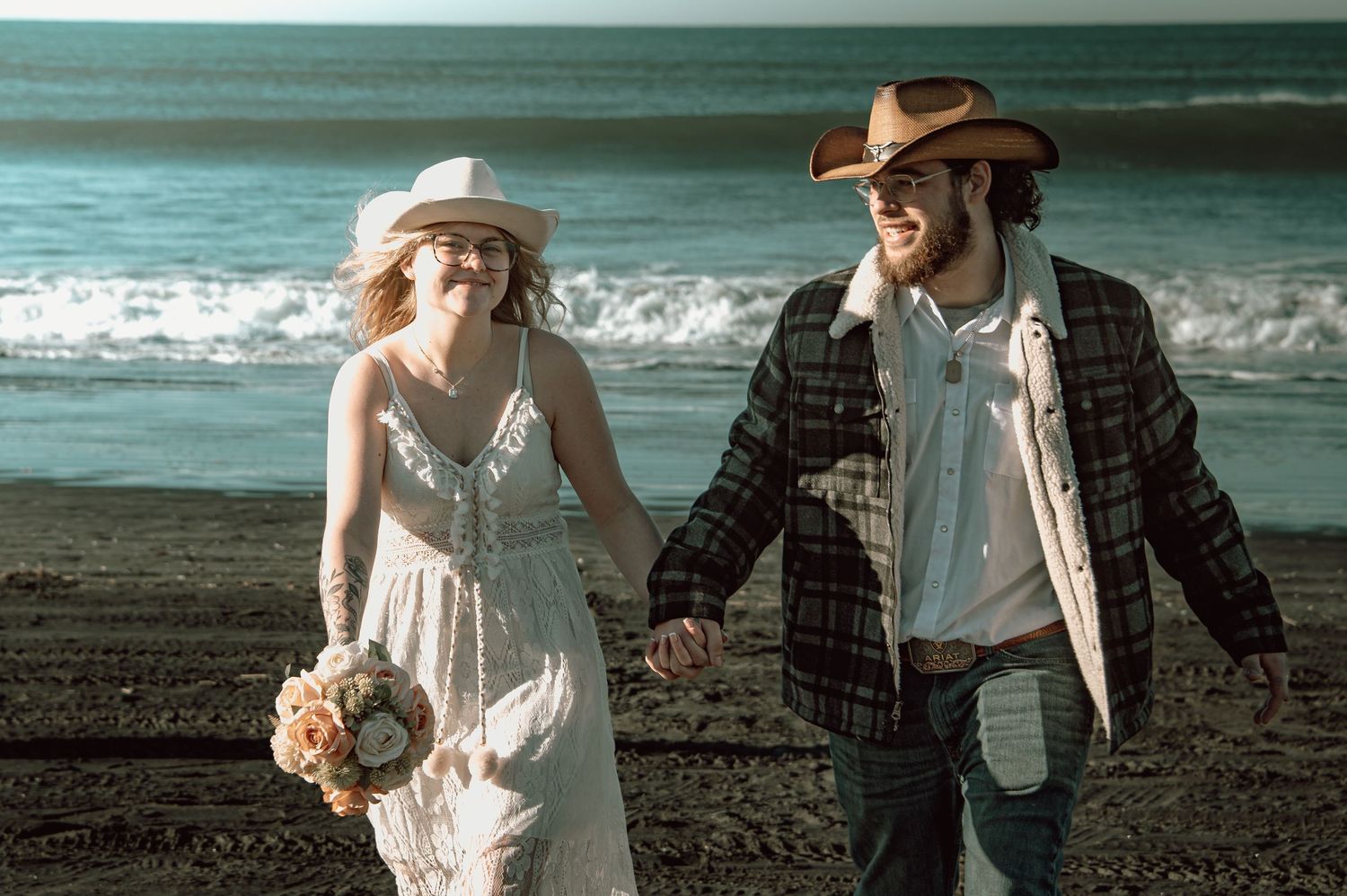 Smiling newlyweds in western hats holding hands and walking on a beach, bride carrying a floral bouquet.