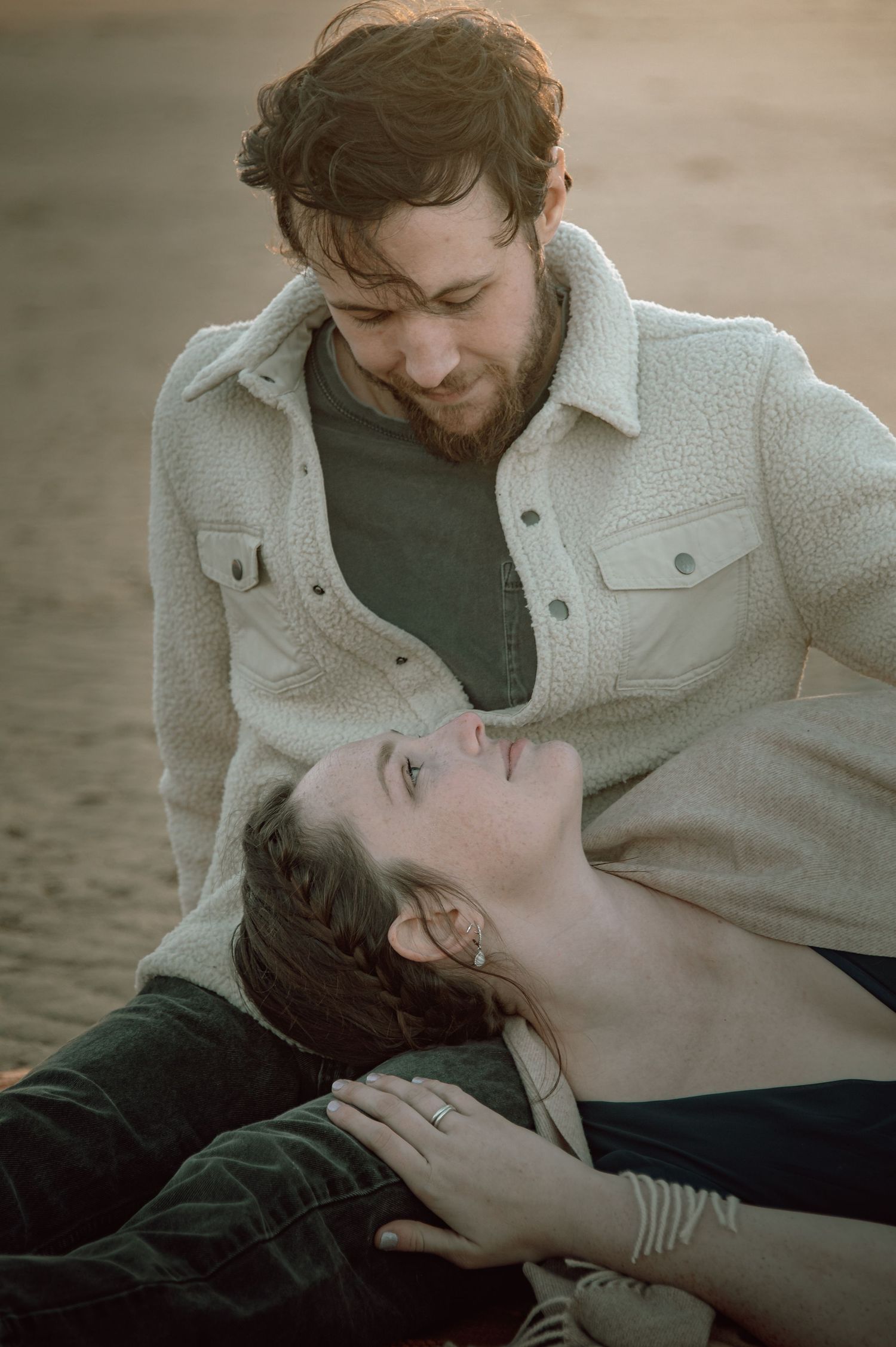 Bearded man in cream jacket gazing lovingly at woman lying across his lap on sandy beach.