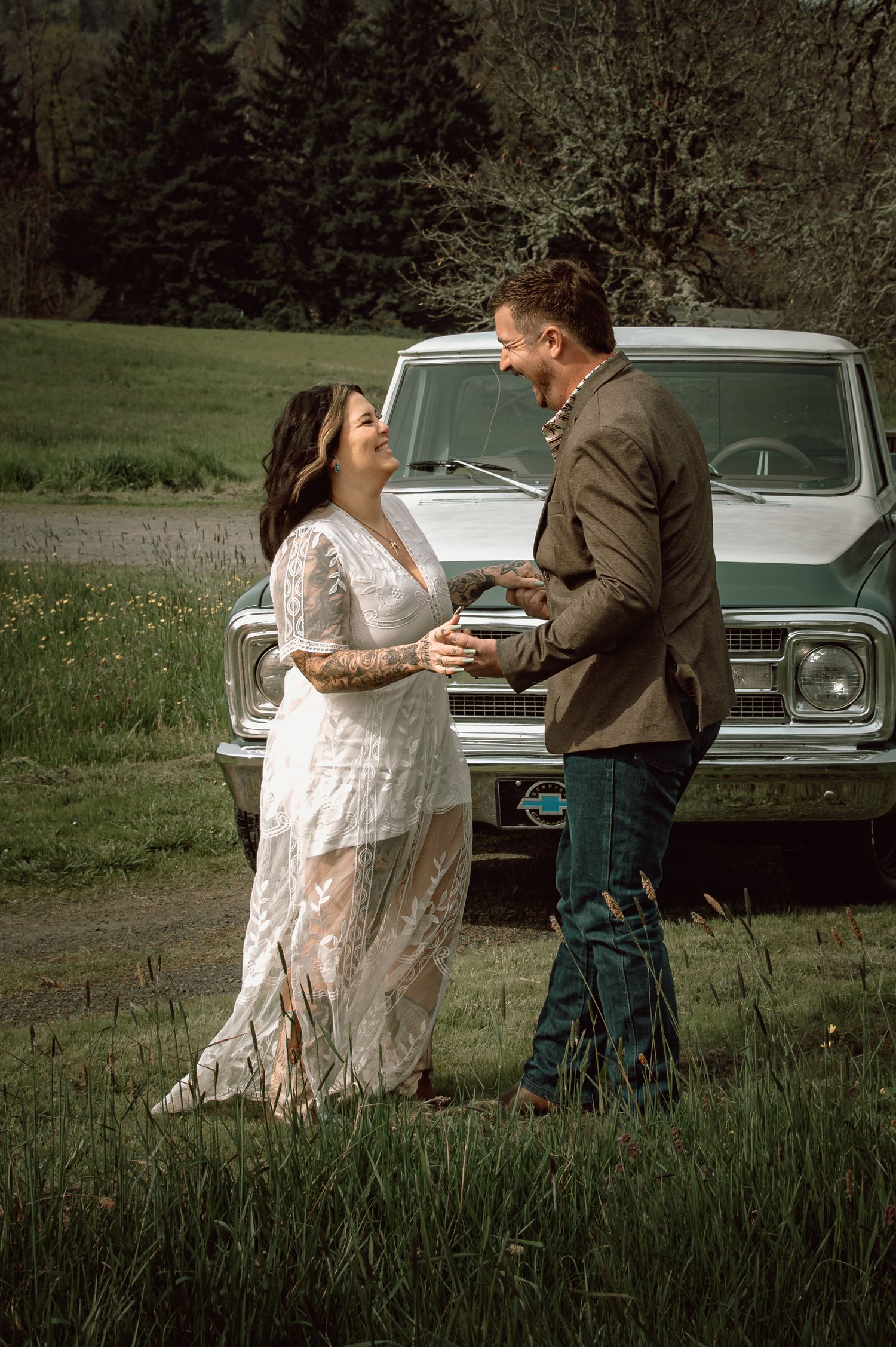 Laughing couple in wedding attire standing before a vintage white truck in a meadow.