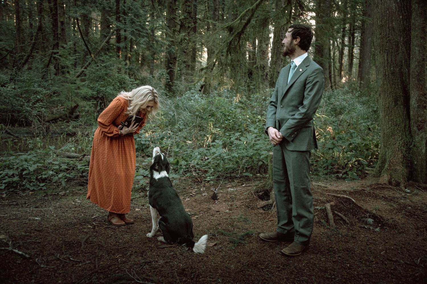 Woman in rust dress and groom in green suit interact with husky dog in forest.