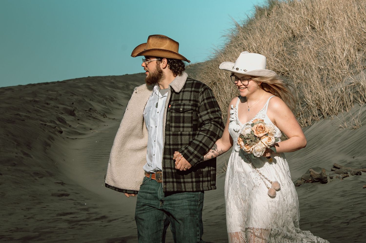 Bride and groom in cowboy hats walking together through sandy coastal dunes with dry beach grass.