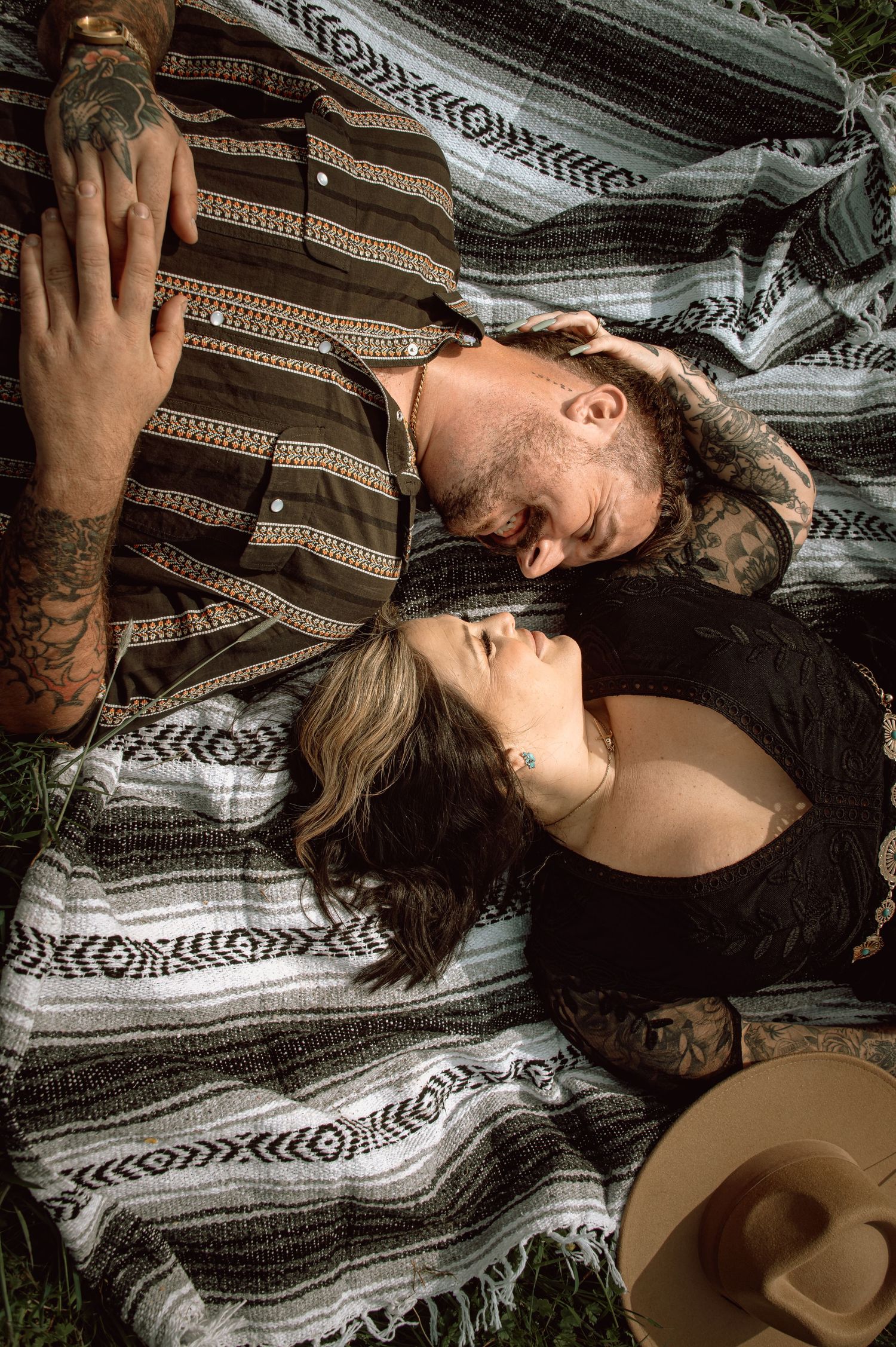 Tattooed couple lying on striped blanket outdoors, sharing an intimate moment face-to-face.