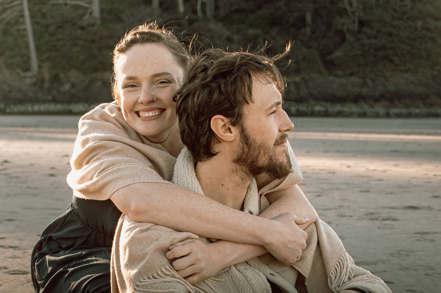 Woman laughing and hugging man from behind on a sandy beach during golden hour.