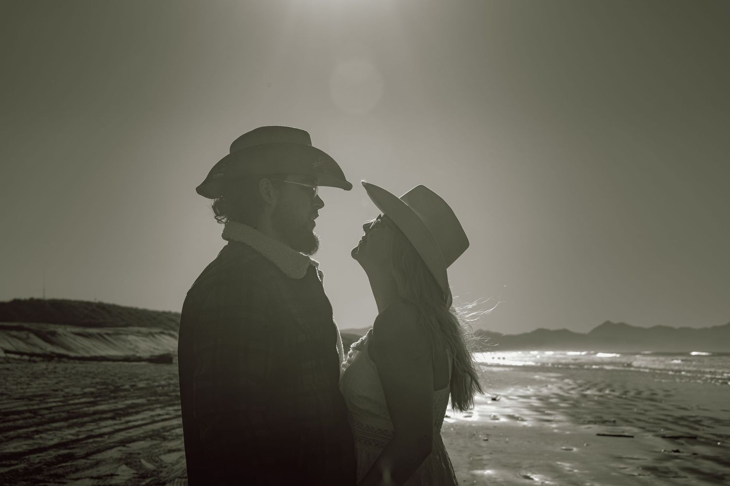 Silhouette of a romantic couple in cowboy hats facing each other on a sunlit beach.