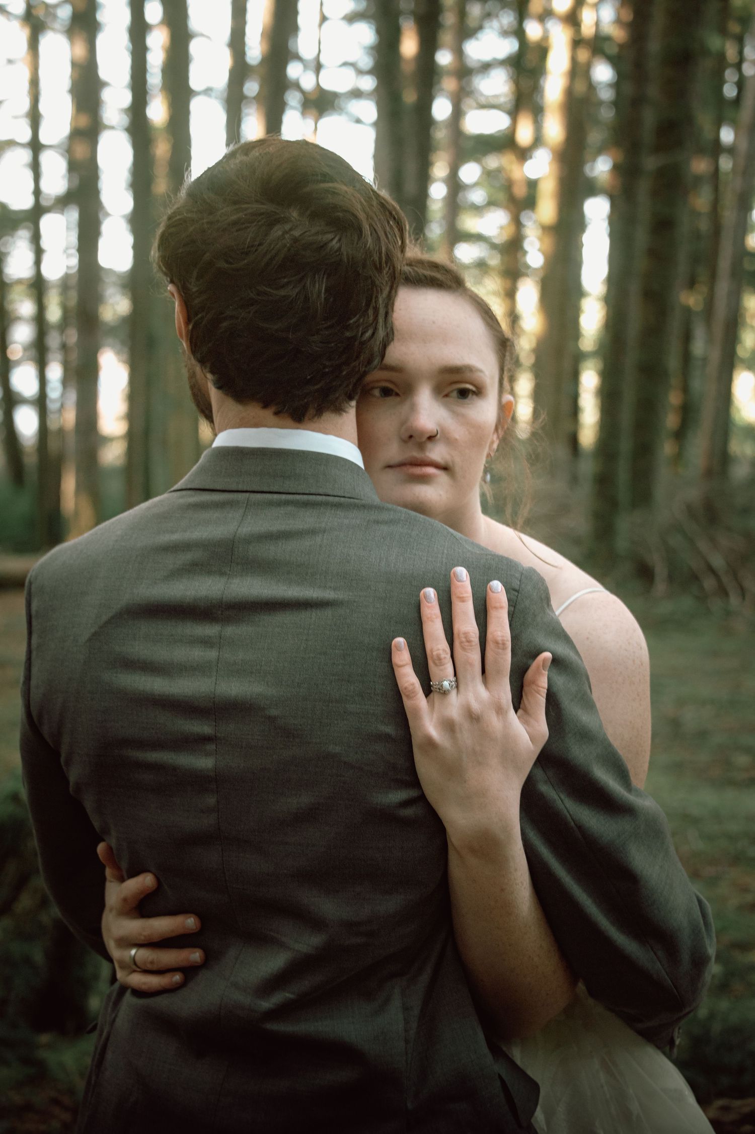 Bride showing diamond ring while embracing groom from behind in sunlit forest portrait.