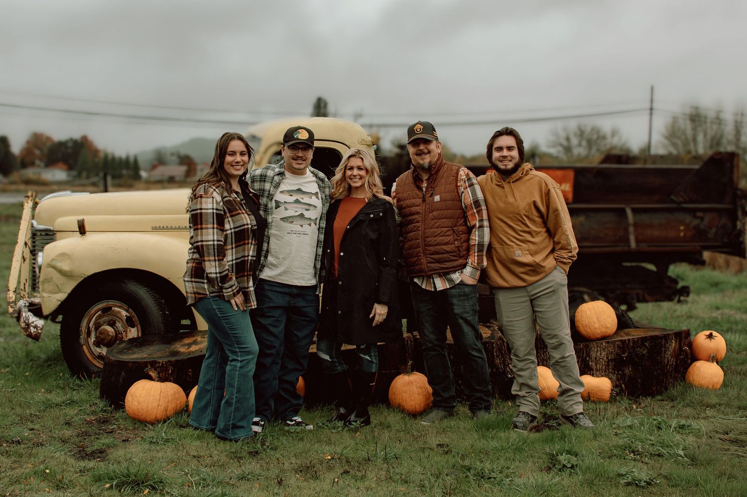 Family of five posing with pumpkins beside a vintage truck in an autumn farm setting.