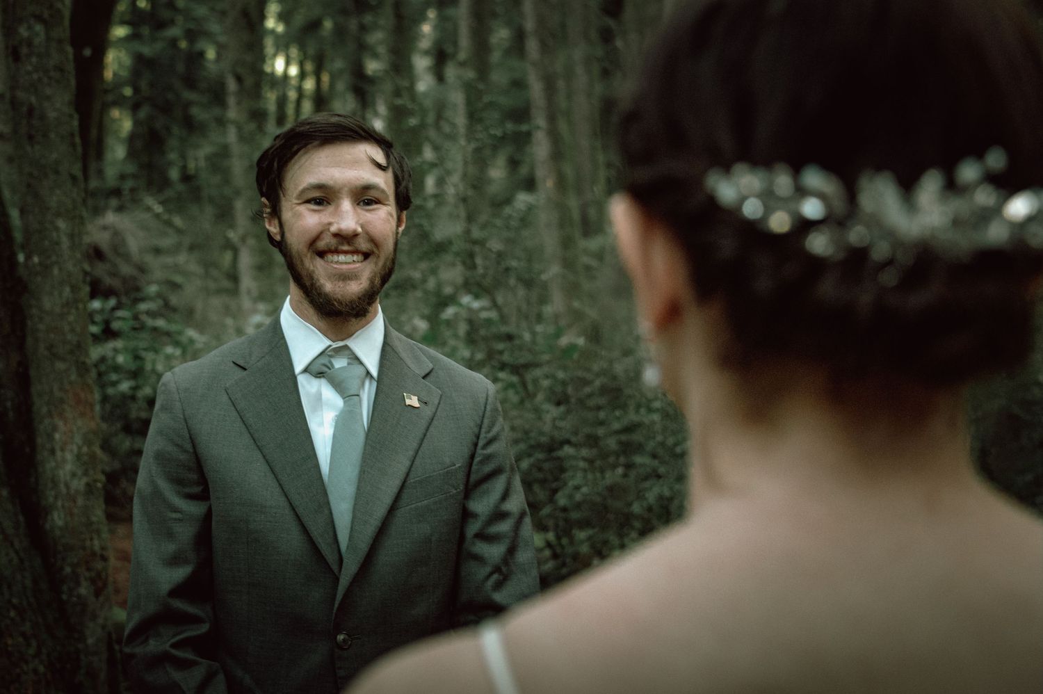 Smiling groom in gray suit reacts with joy seeing his bride during a forest wedding first look.