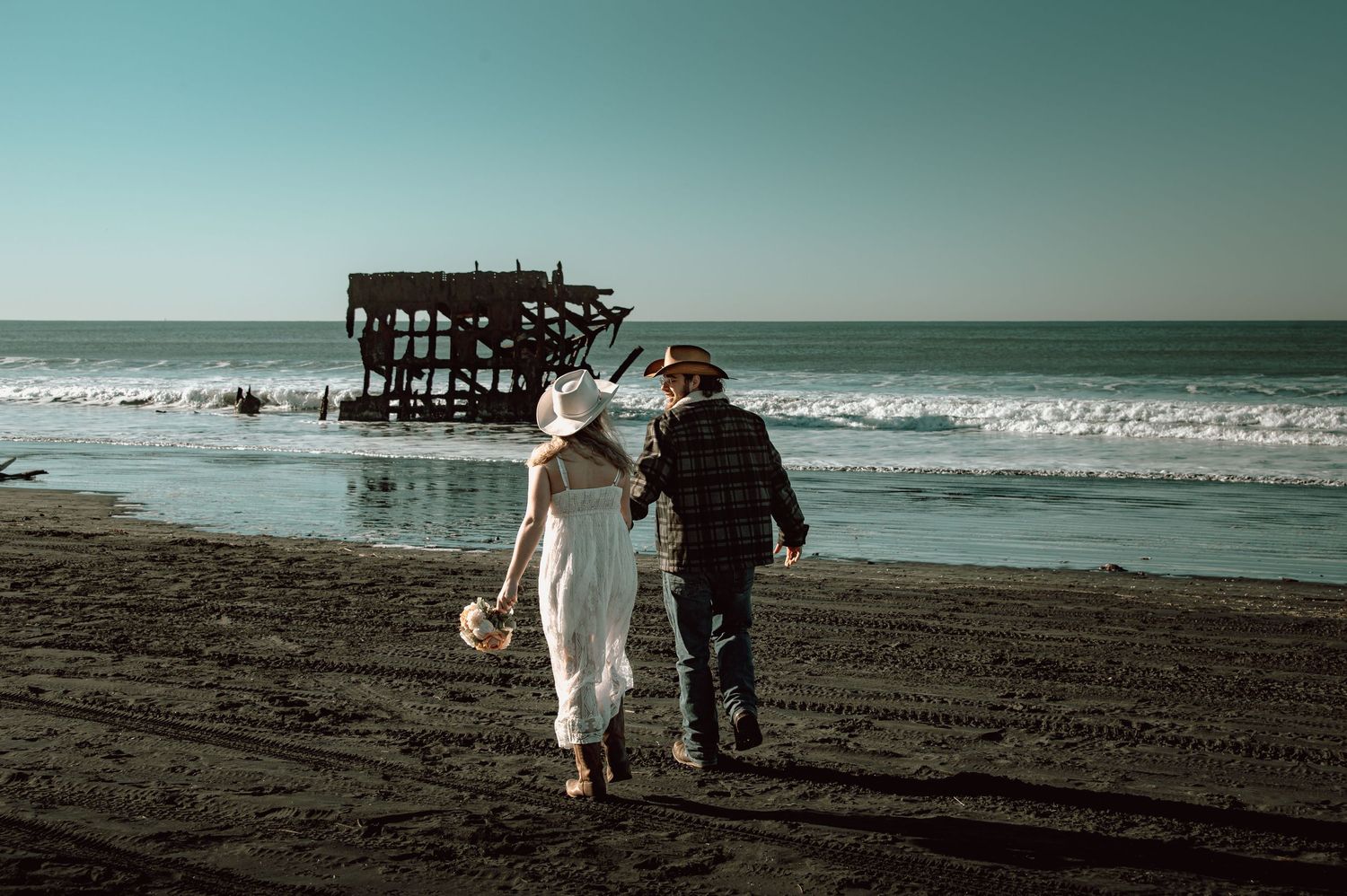 Bride and groom walking toward a historic shipwreck on a dark sand beach under a clear blue sky.