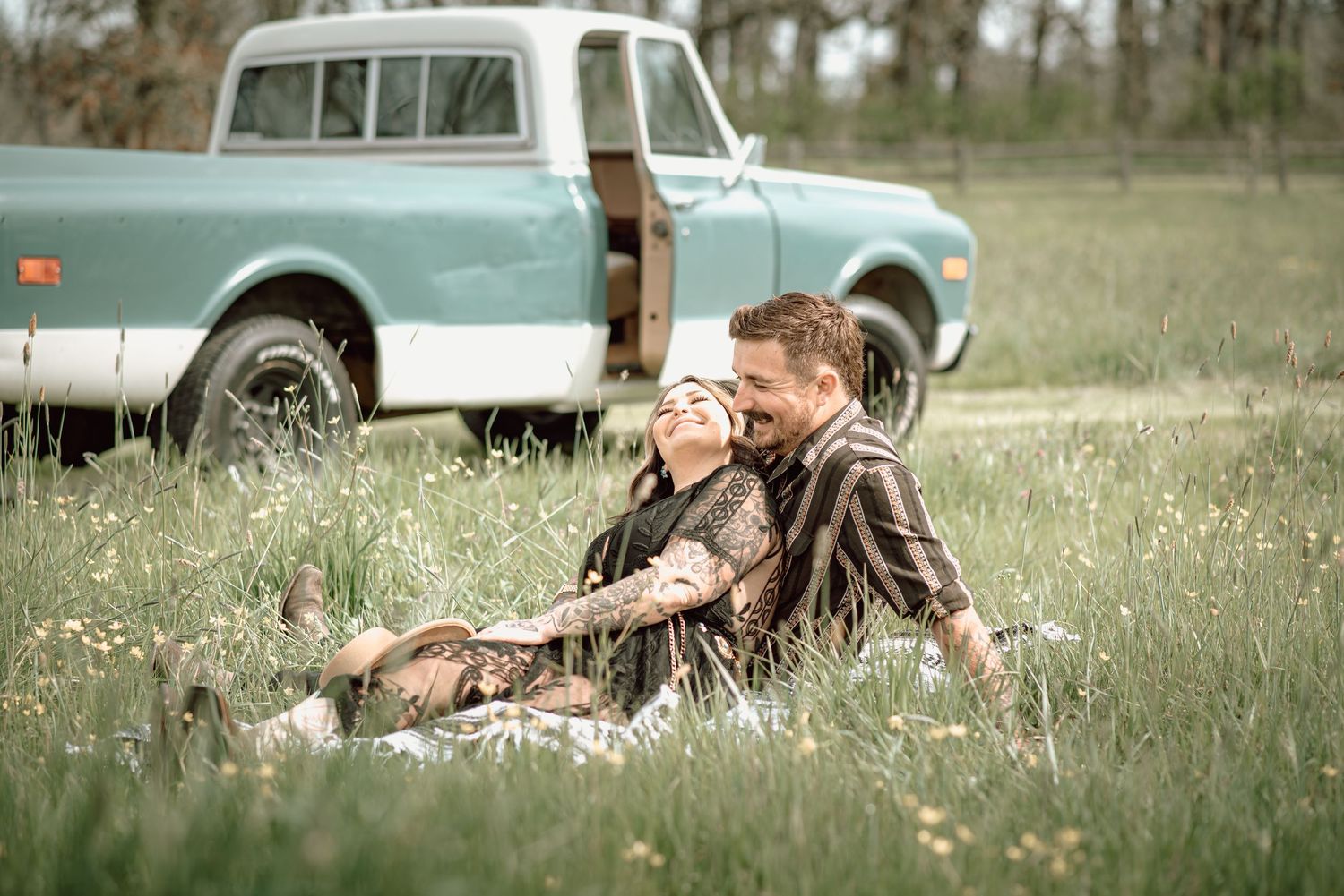 Tattooed couple laughing together in a wildflower field beside a classic teal pickup truck.