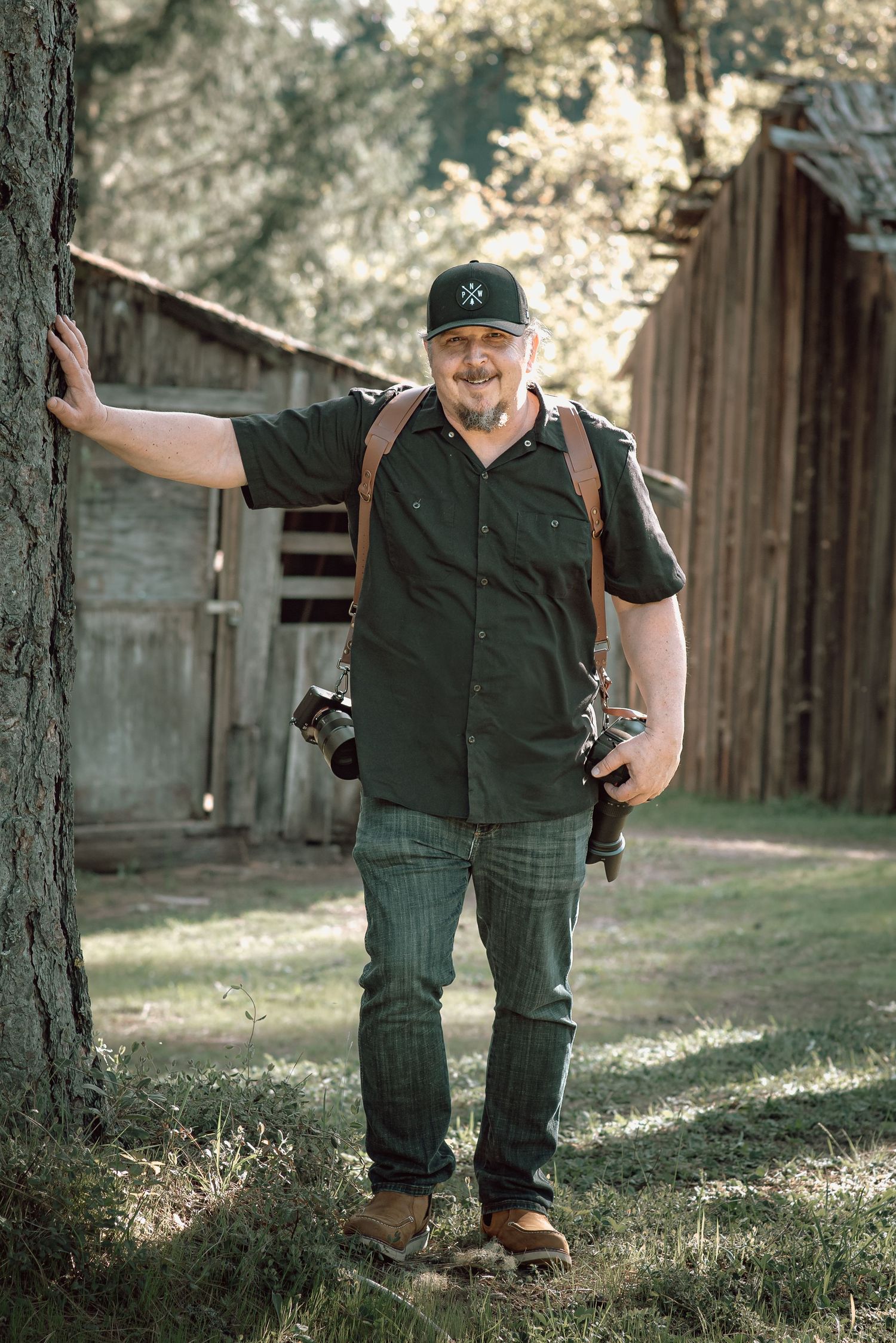 Bearded male photographer with cameras leaning against a tree near a rustic wooden barn.