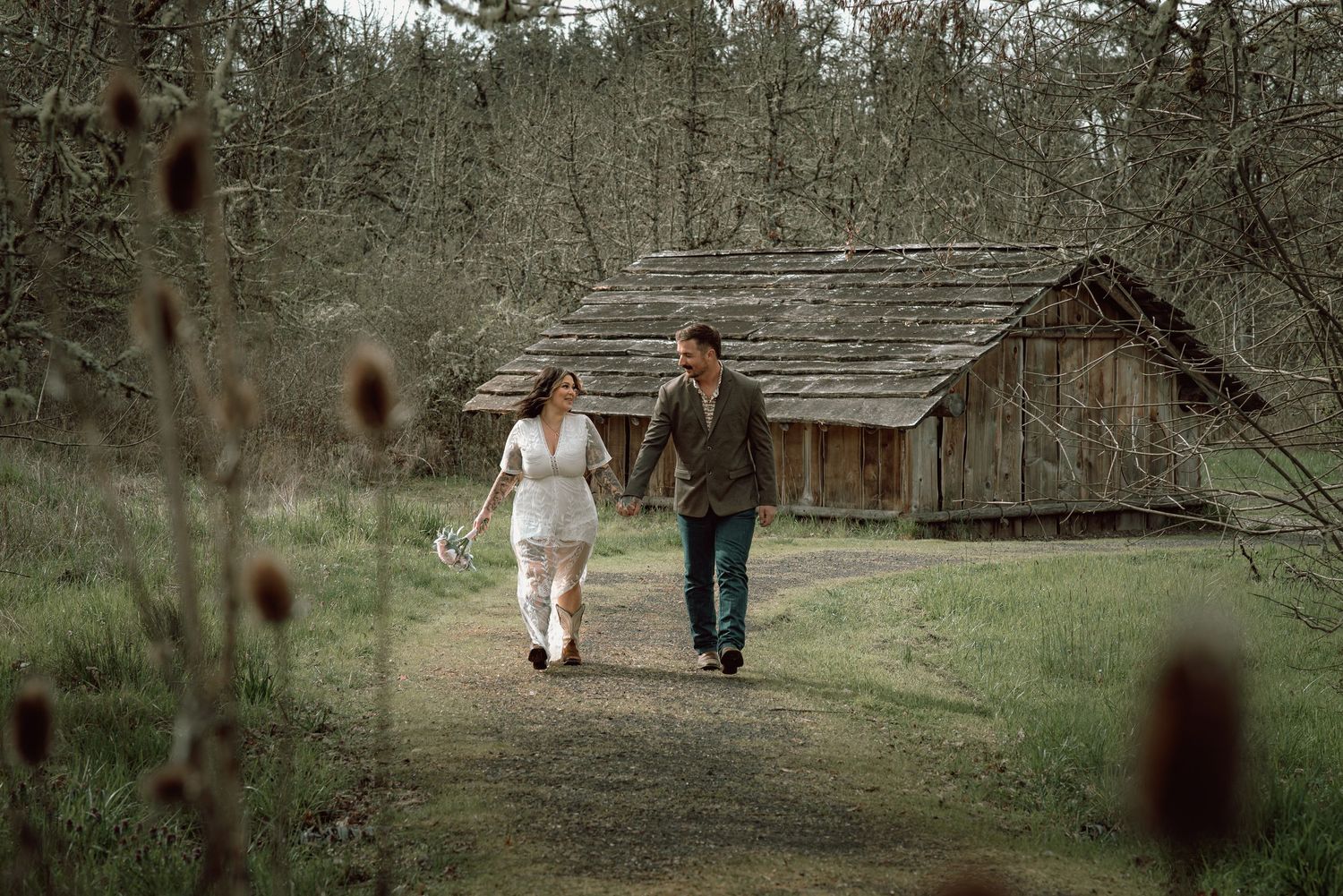 Newlywed couple walking hand in hand on a dirt path near a rustic wooden barn.