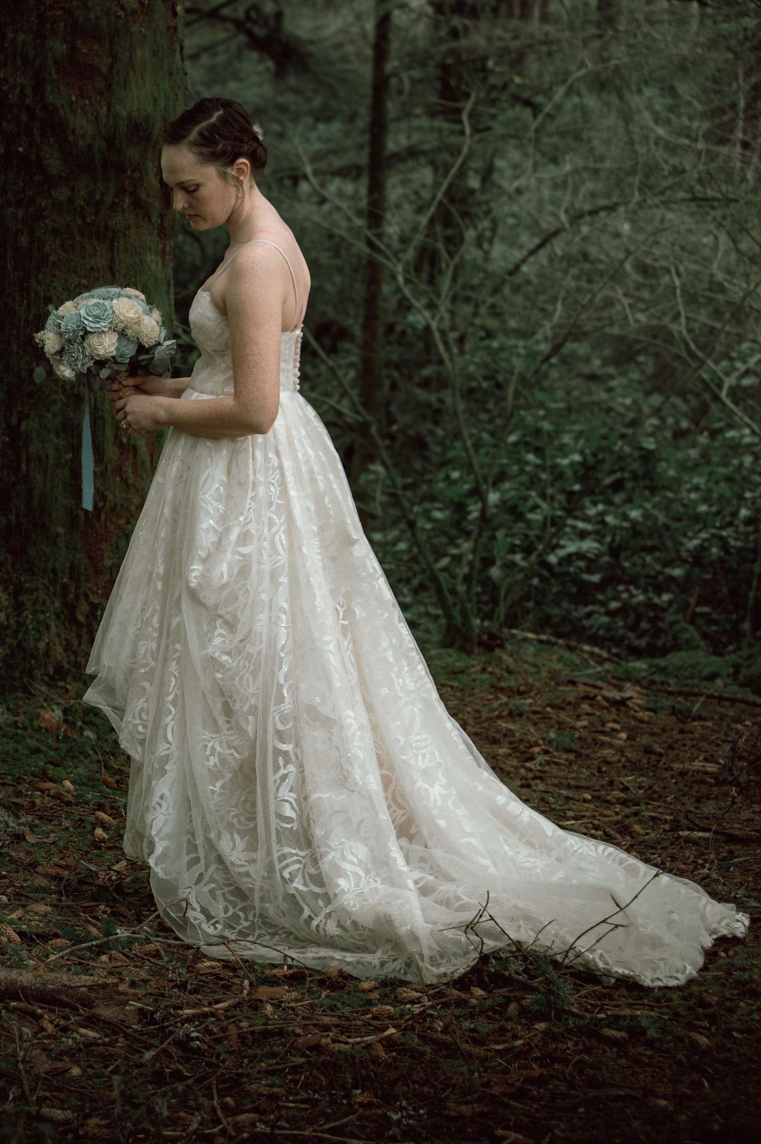 Bride in lace wedding gown holding bouquet while standing among trees in a forest setting.