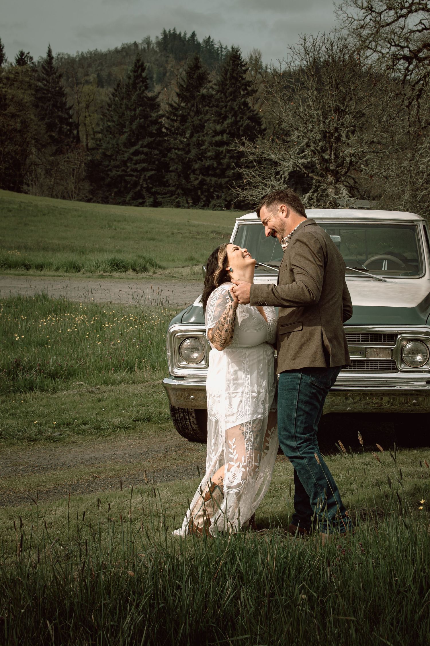 Couple dancing romantically in a green field beside a vintage truck with forested hills in the background.