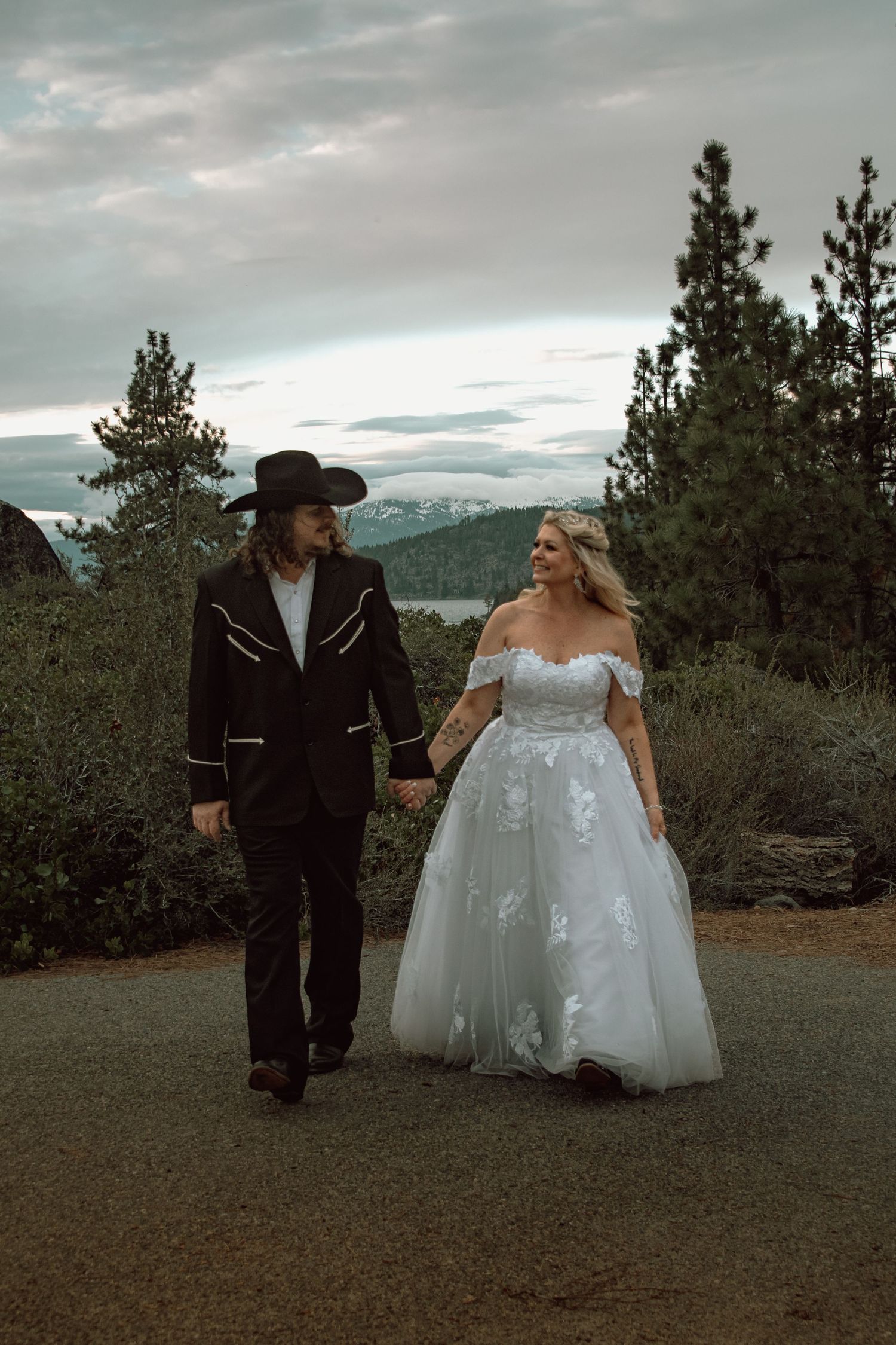 Bride in floral ballgown and groom in black cowboy hat strolling along scenic mountain path.