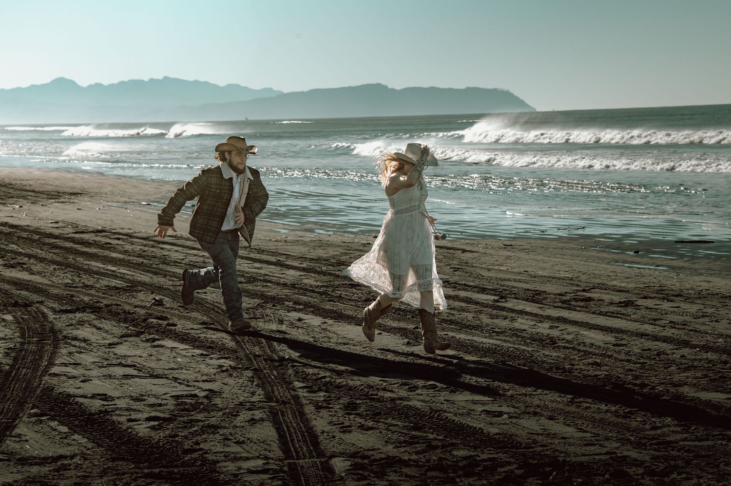 Joyful couple running along sandy beach with ocean waves, bride in flowing white dress.