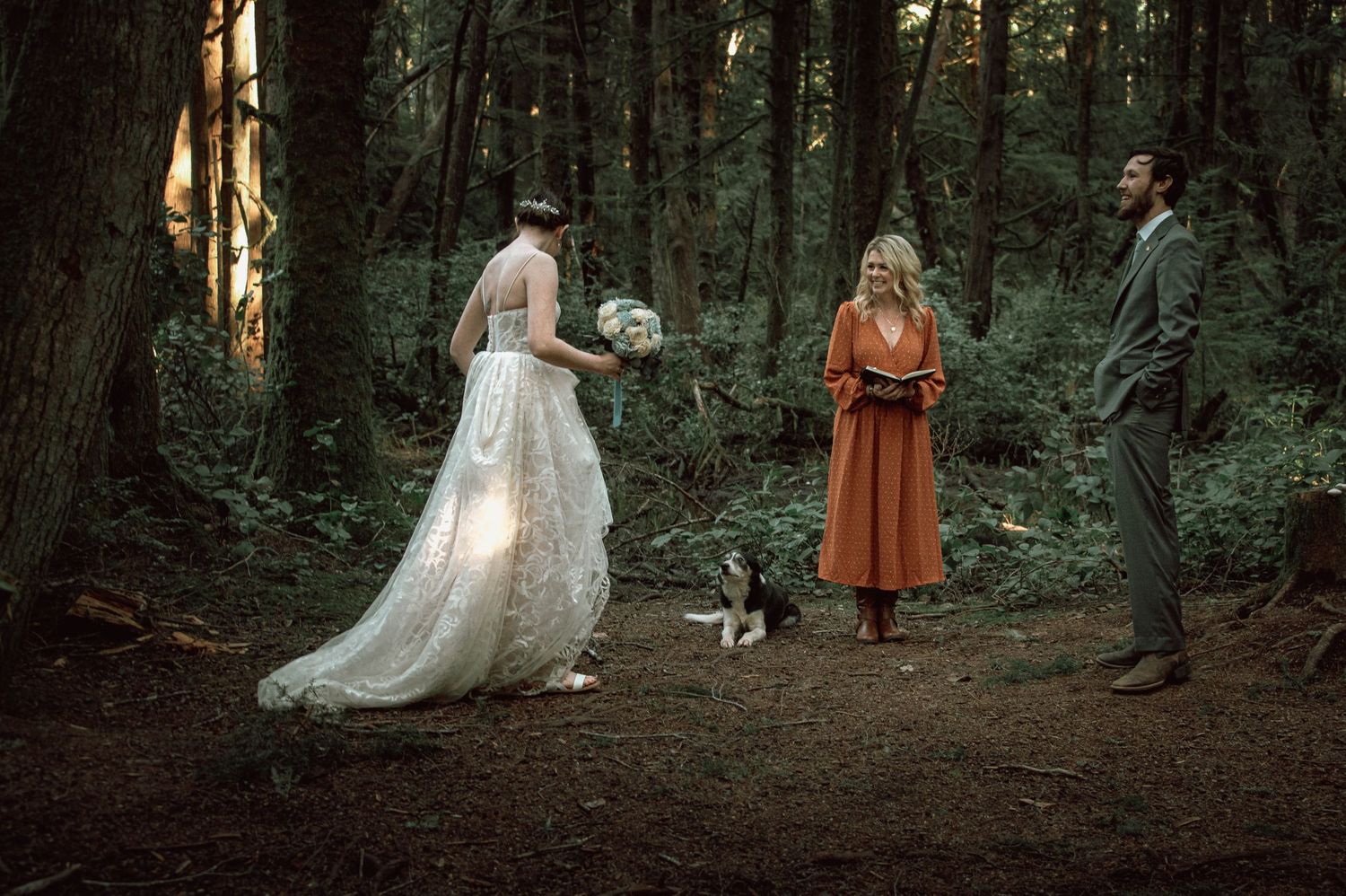 Bride in white lace gown meets groom and officiant in orange dress with a dog during forest ceremony.