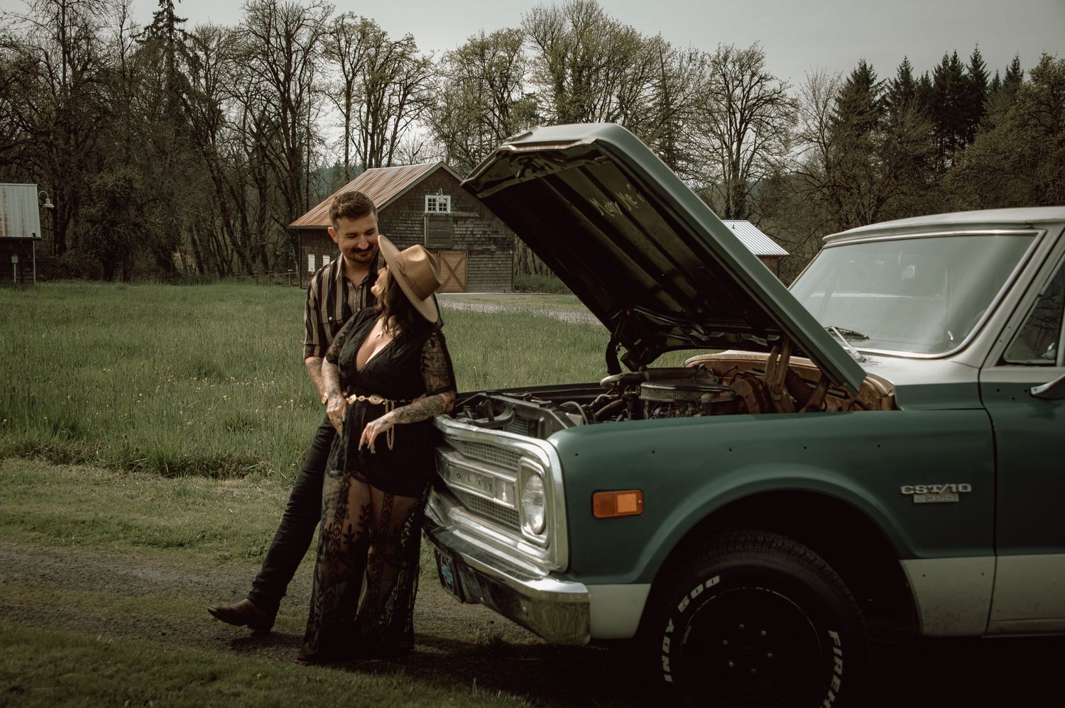 Tattooed man leaning against a vintage teal truck with open hood in a rural green field setting.