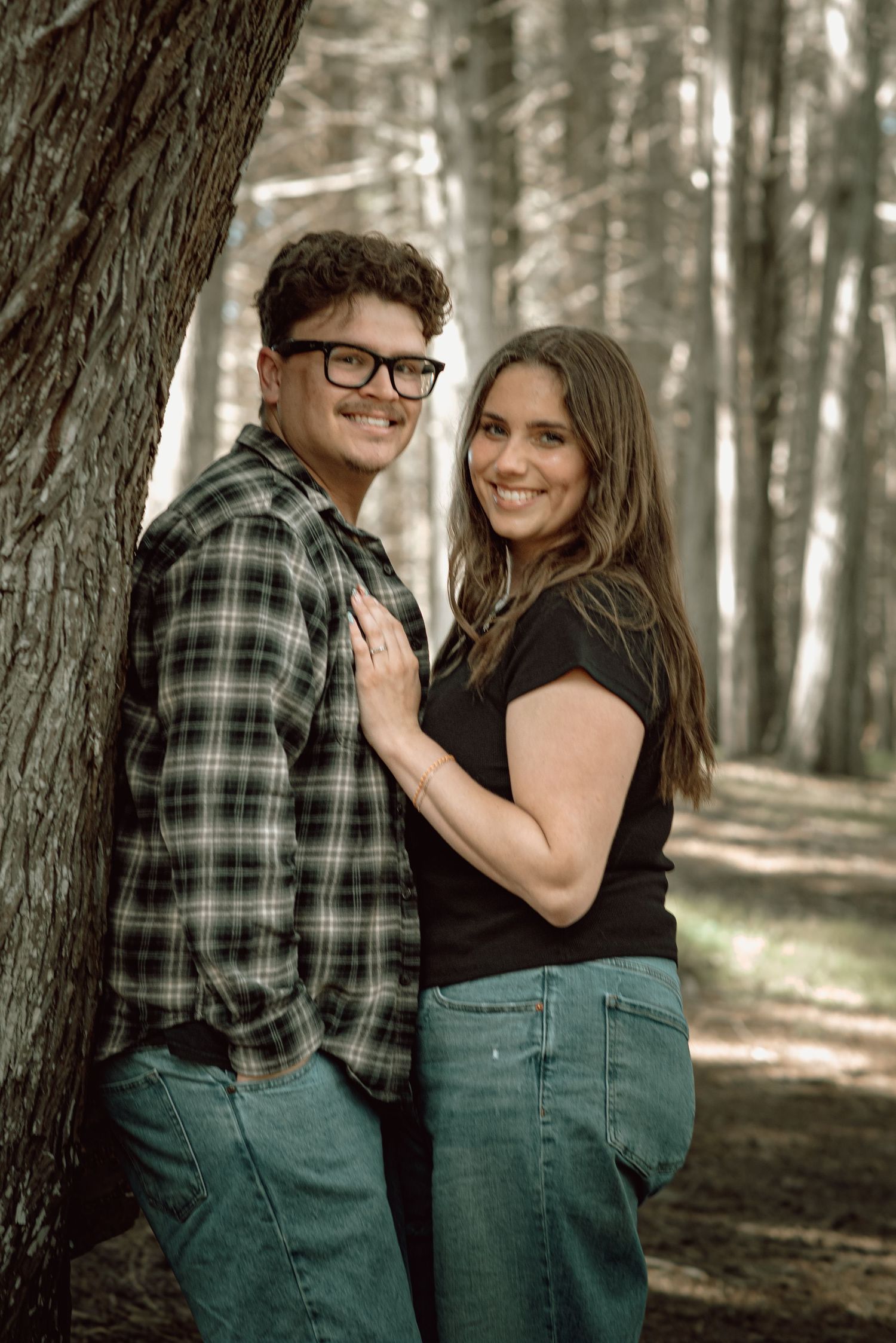 Smiling couple poses together against a tree trunk in a peaceful forest setting.