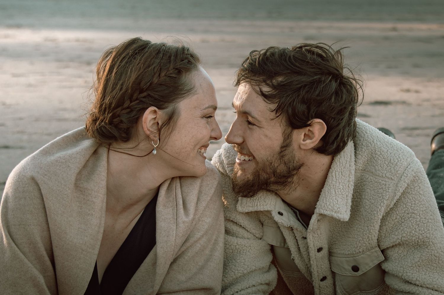 Smiling couple sitting close together on beach, gazing at each other during golden hour.