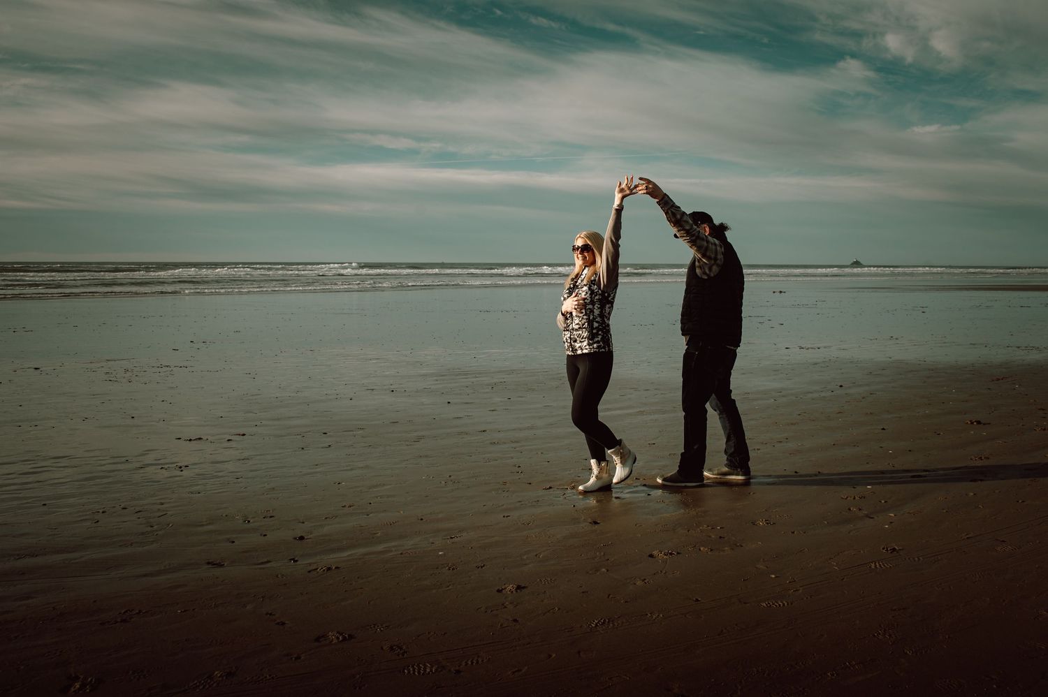 Couple dancing on a sandy beach with ocean waves and dramatic cloudy sky behind them.