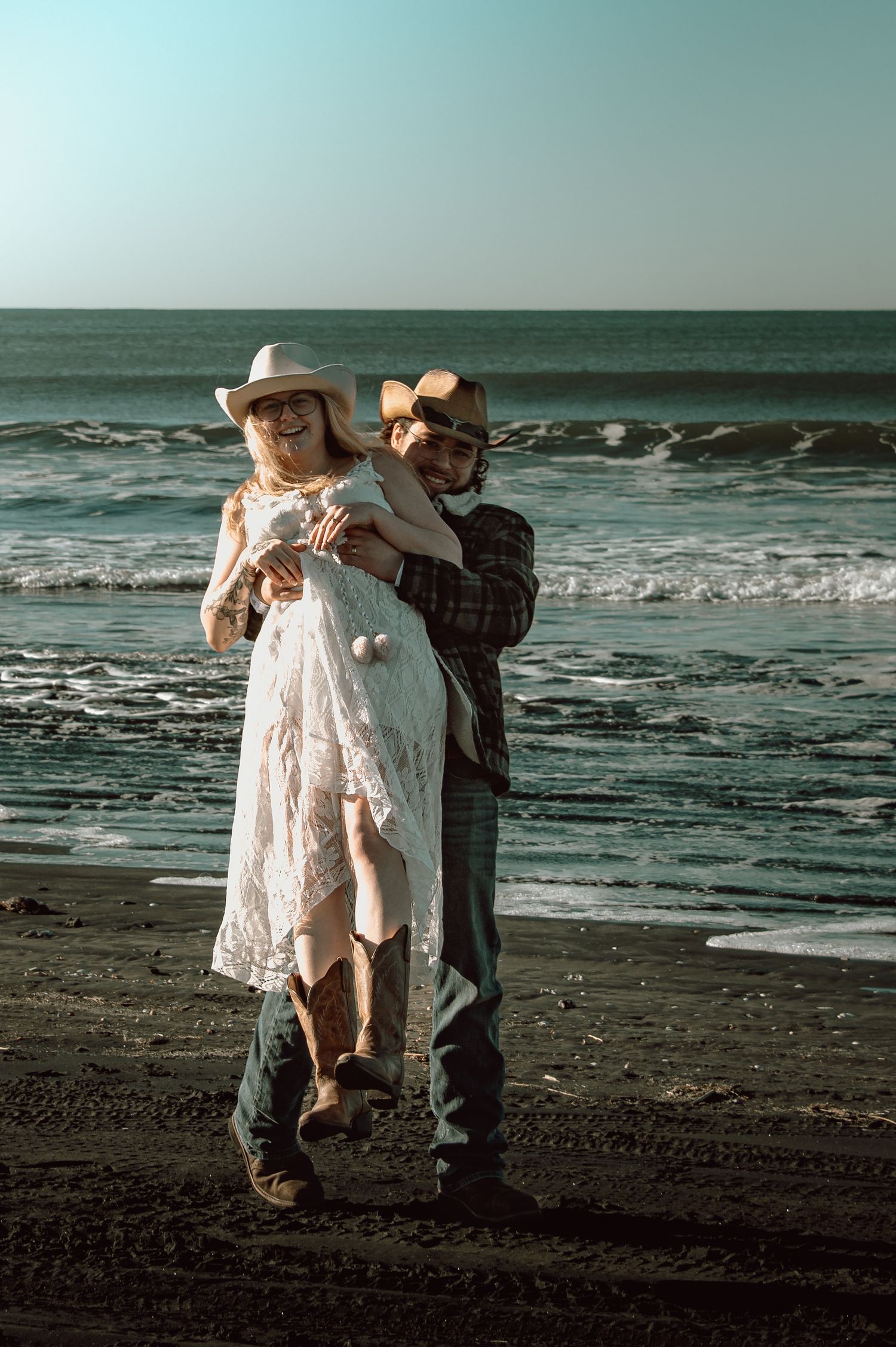 Couple in cowboy hats cuddle together on dark sand beach with ocean backdrop.