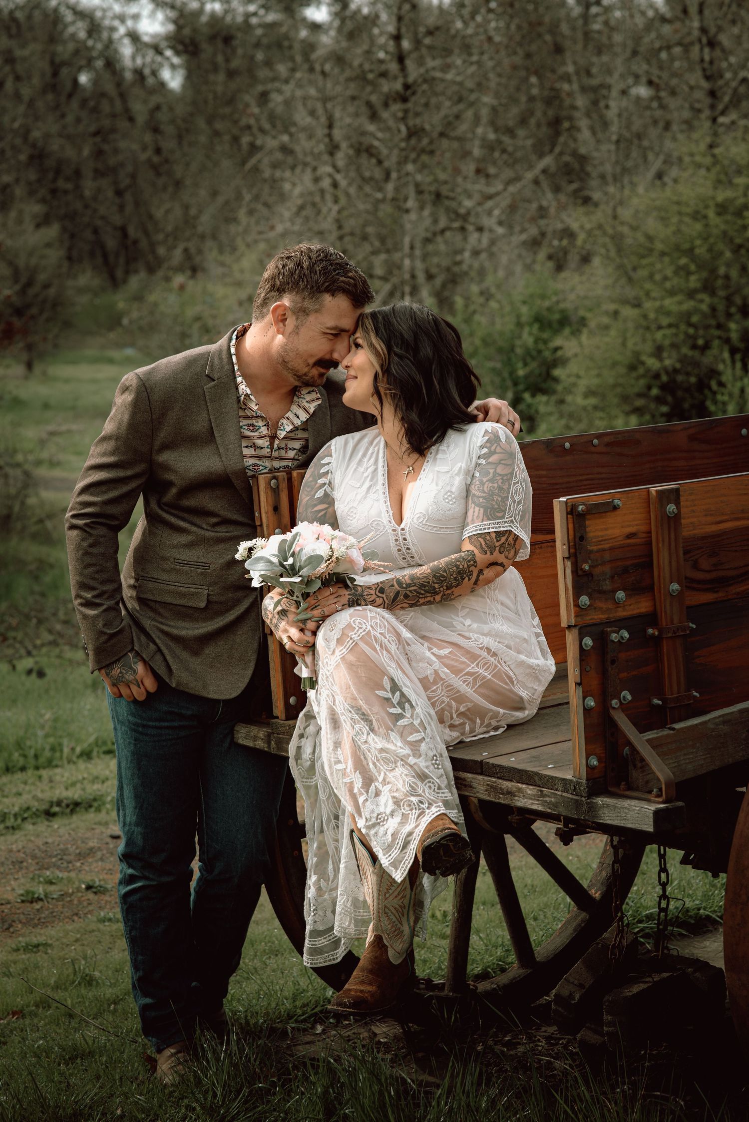 Tattooed bride in lace dress sitting on wooden wagon with groom in olive blazer outdoors.