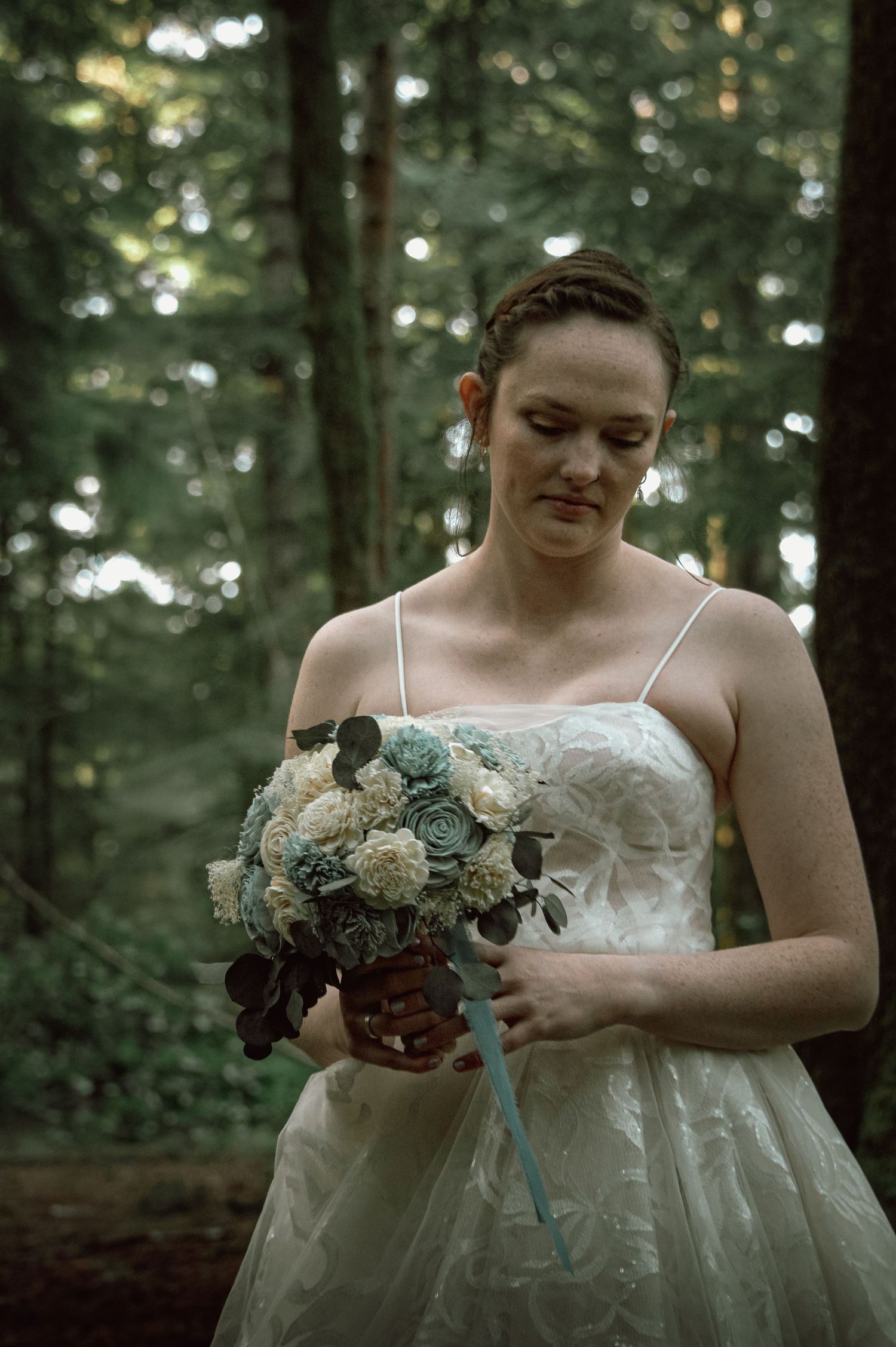Bride in white lace gown holding blue and white floral bouquet, looking down in a lush forest setting.