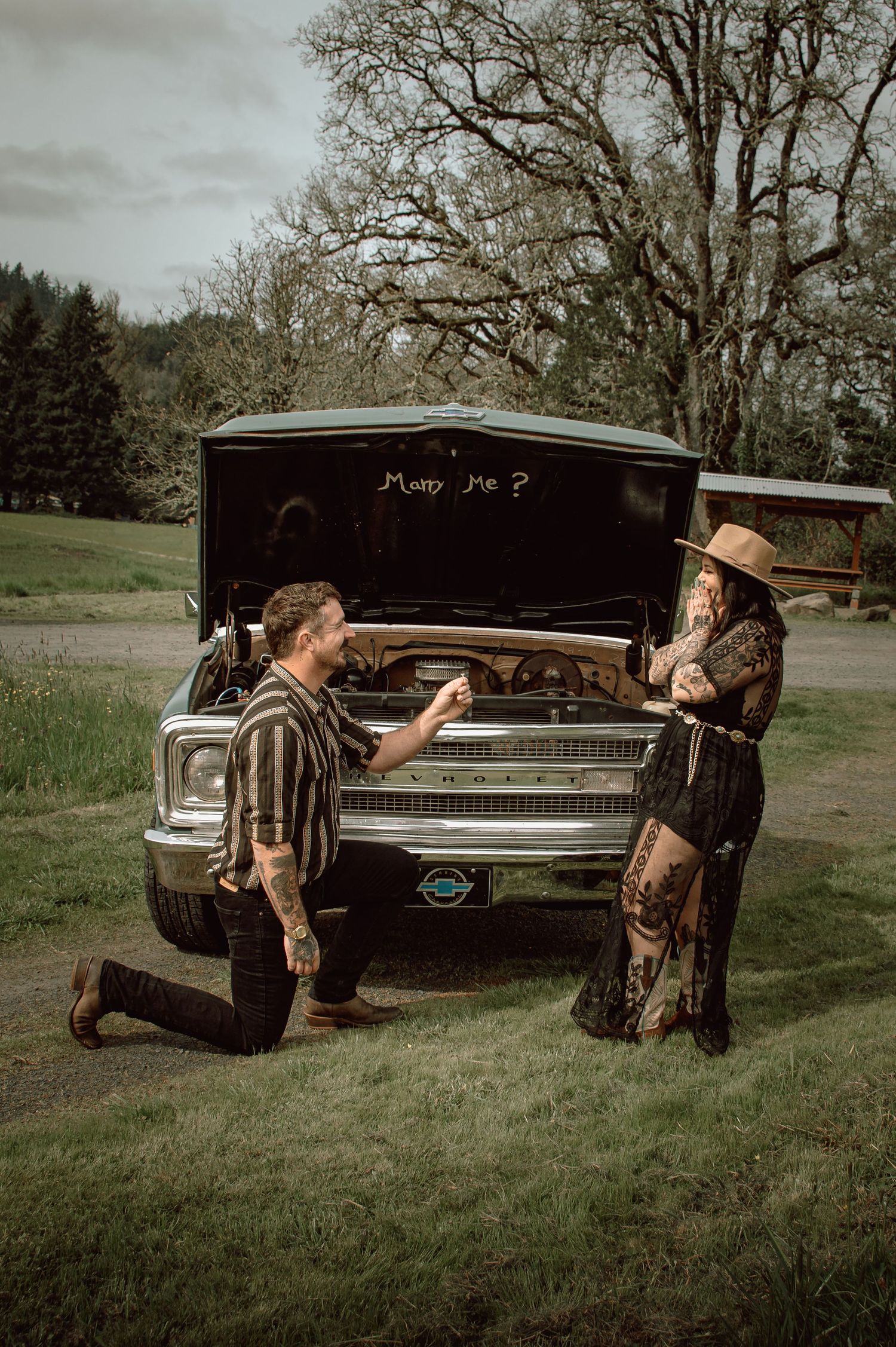 Man proposing on one knee beside a vintage truck with hood open reading Marry Me in a scenic field.