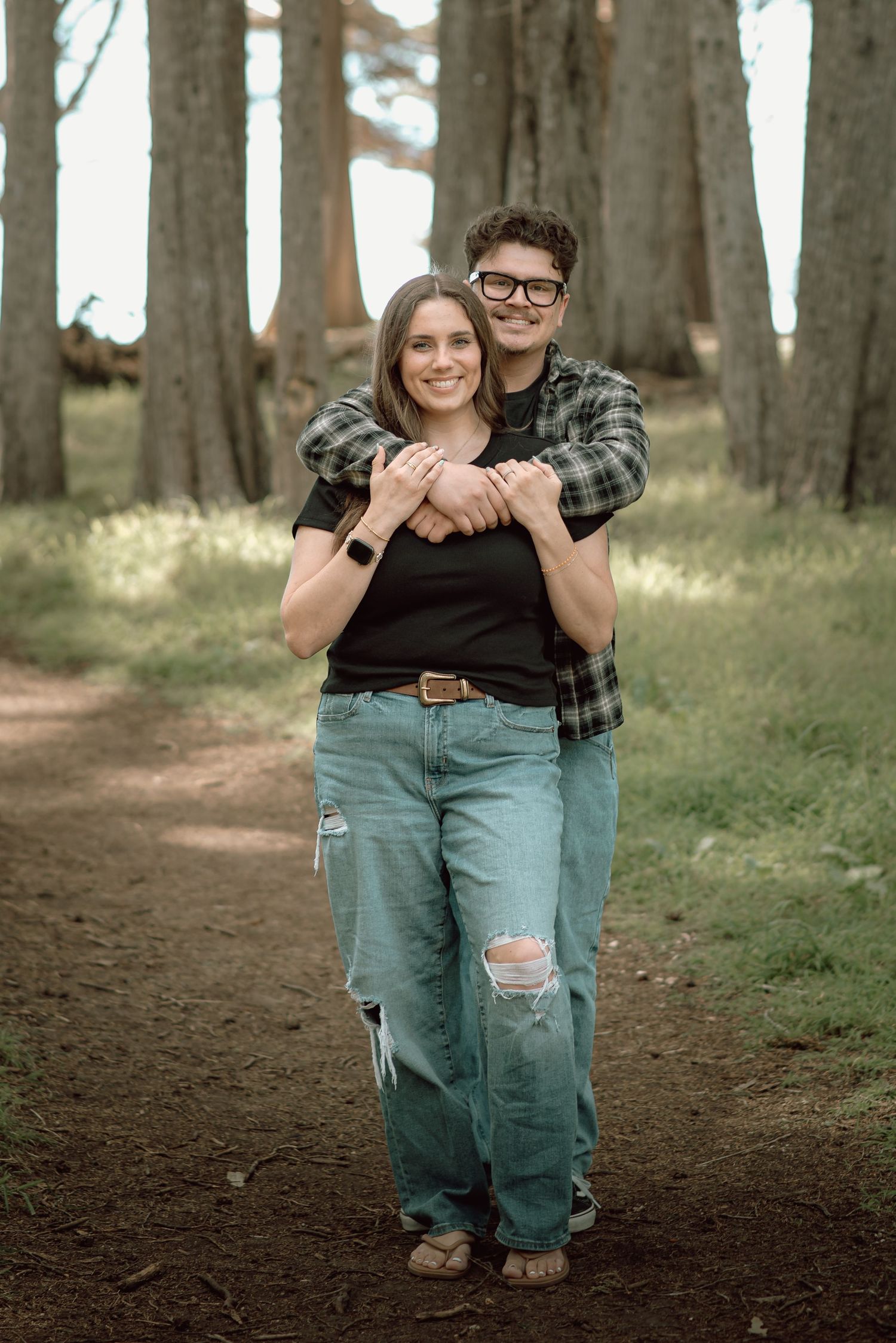 Man hugging woman from behind on a forest path, both smiling, wearing casual jeans and plaid shirt.