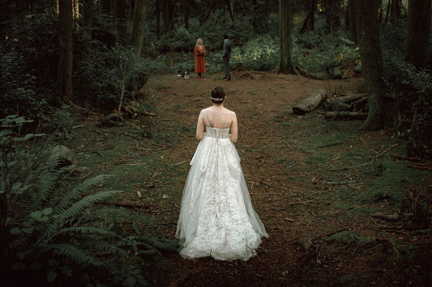 Bride in elegant white floral gown walks through lush green forest toward officiant.
