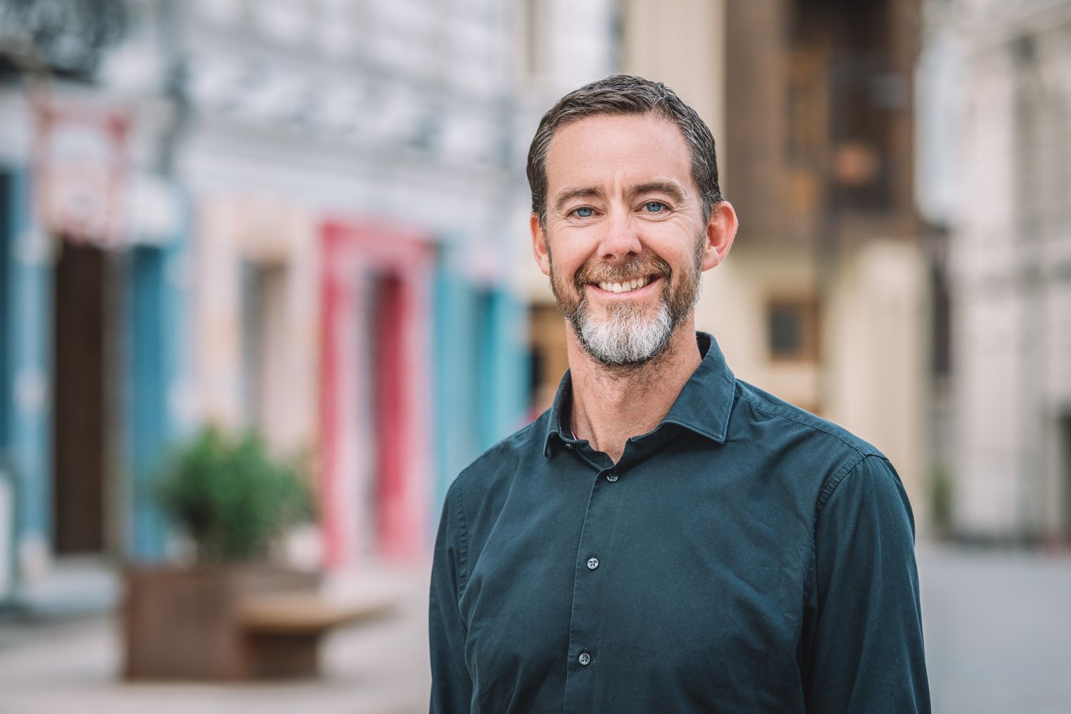 Man in a dark shirt standing confidently in a lively, urban outdoor setting.