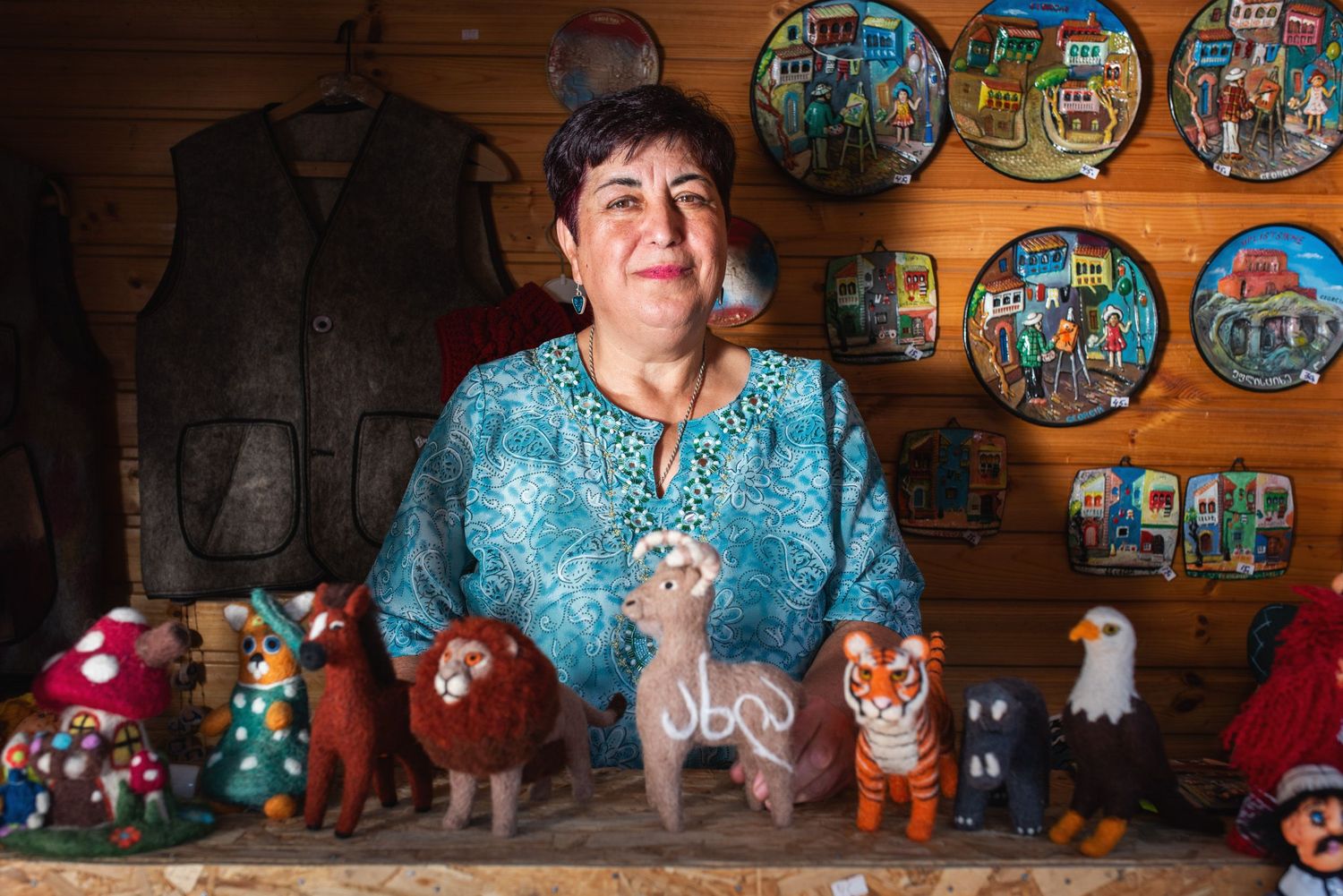 Elderly woman presenting hand-painted ceramic items in a traditional Georgian interior.