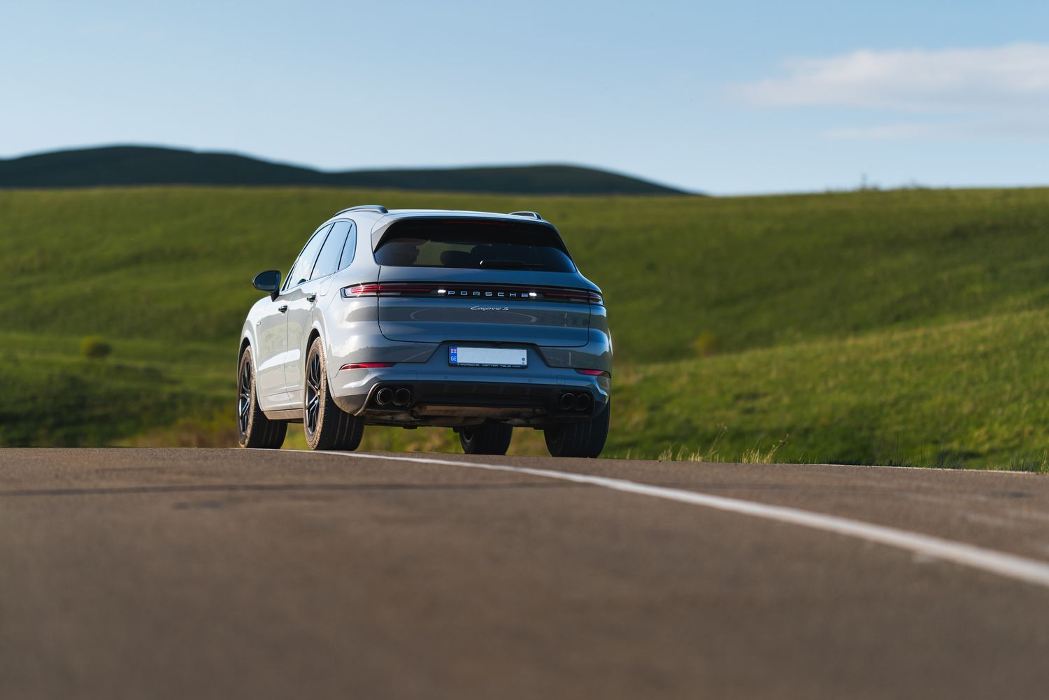 White SUV driving down a rural asphalt road surrounded by green rolling hills.