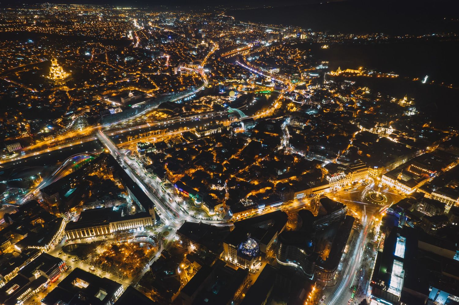 Drone view of a brightly lit cityscape at night with intersecting roads and traffic flow.