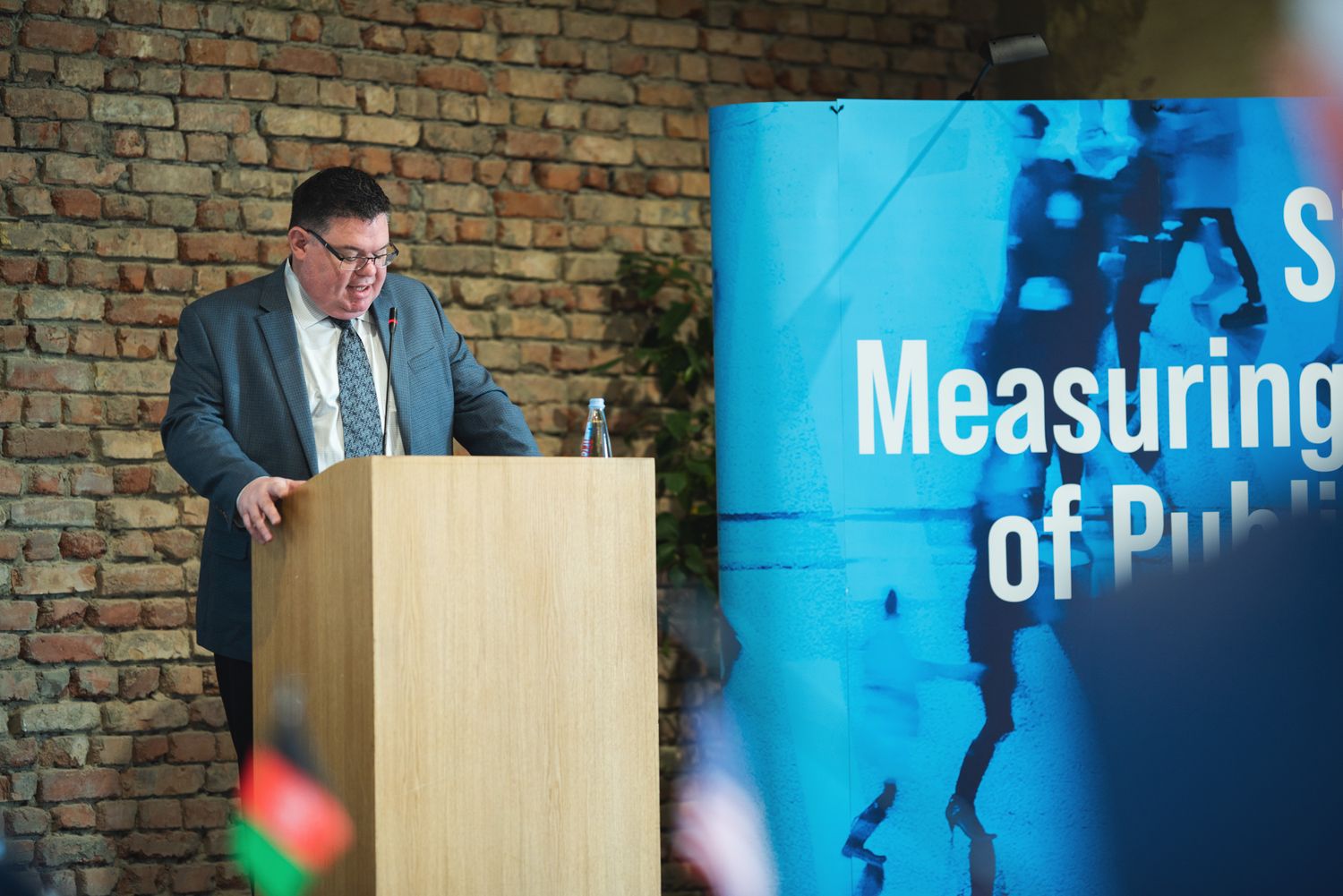 Man giving a talk on stage beside a large screen displaying “Measuring…” and blue-themed visuals.