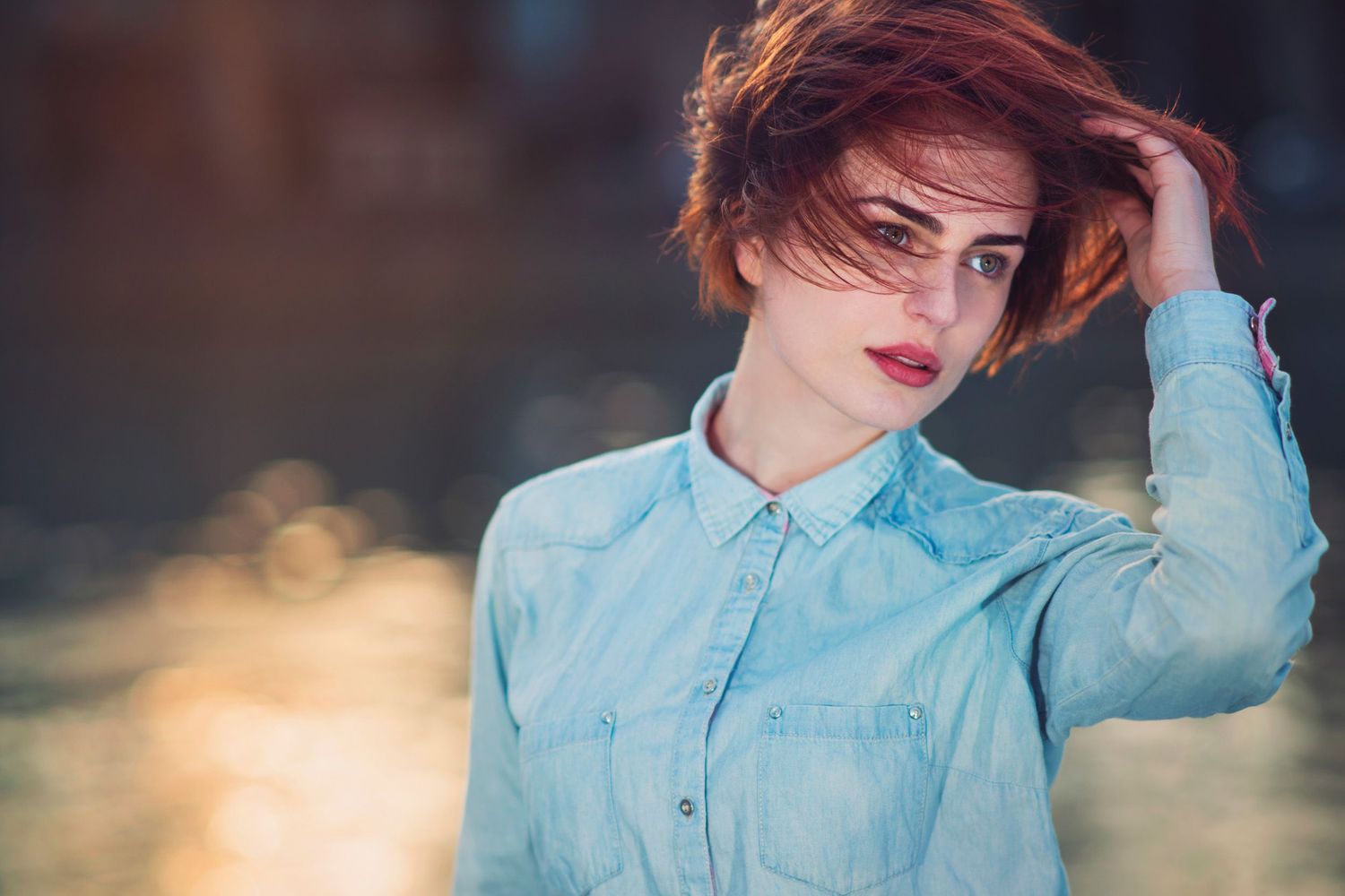 Stylish woman in a denim shirt posing with her hand in hair — soft backlight and shallow depth of field.