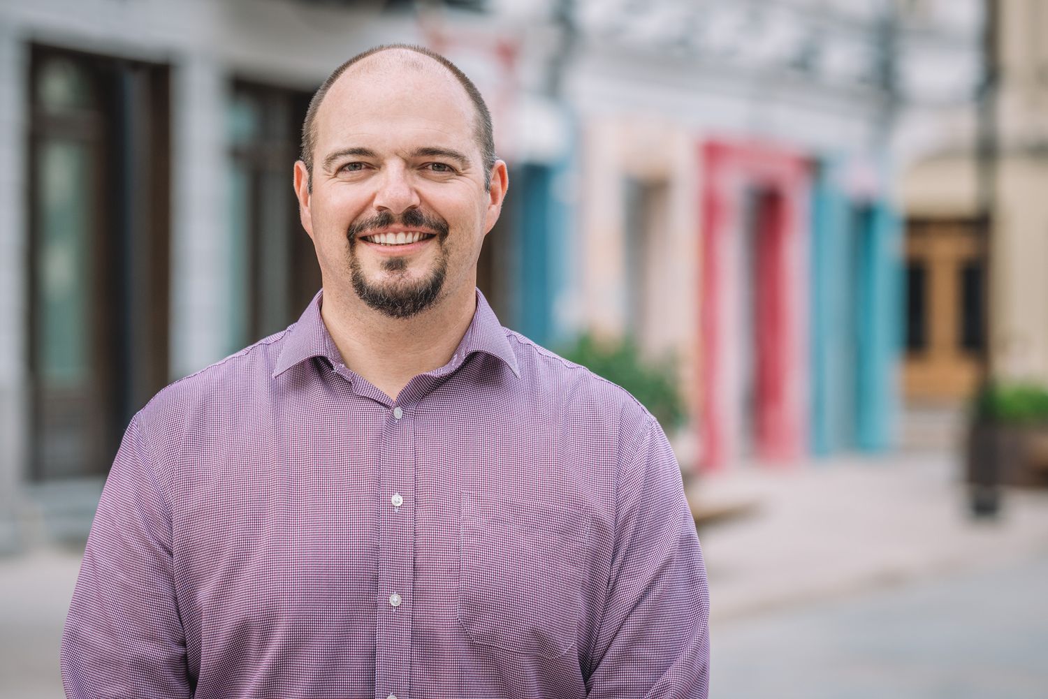 Bald man with a beard smiling in front of pastel-colored buildings on a city street.